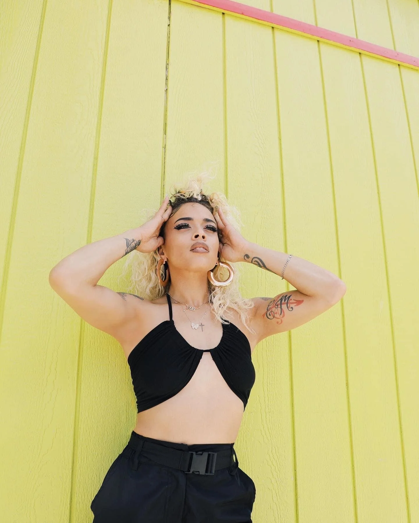 A woman with curly blonde hair wearing a black crop top and black pants, standing against a yellow wall with pink trim, holding her head with both hands and looking into the distance.