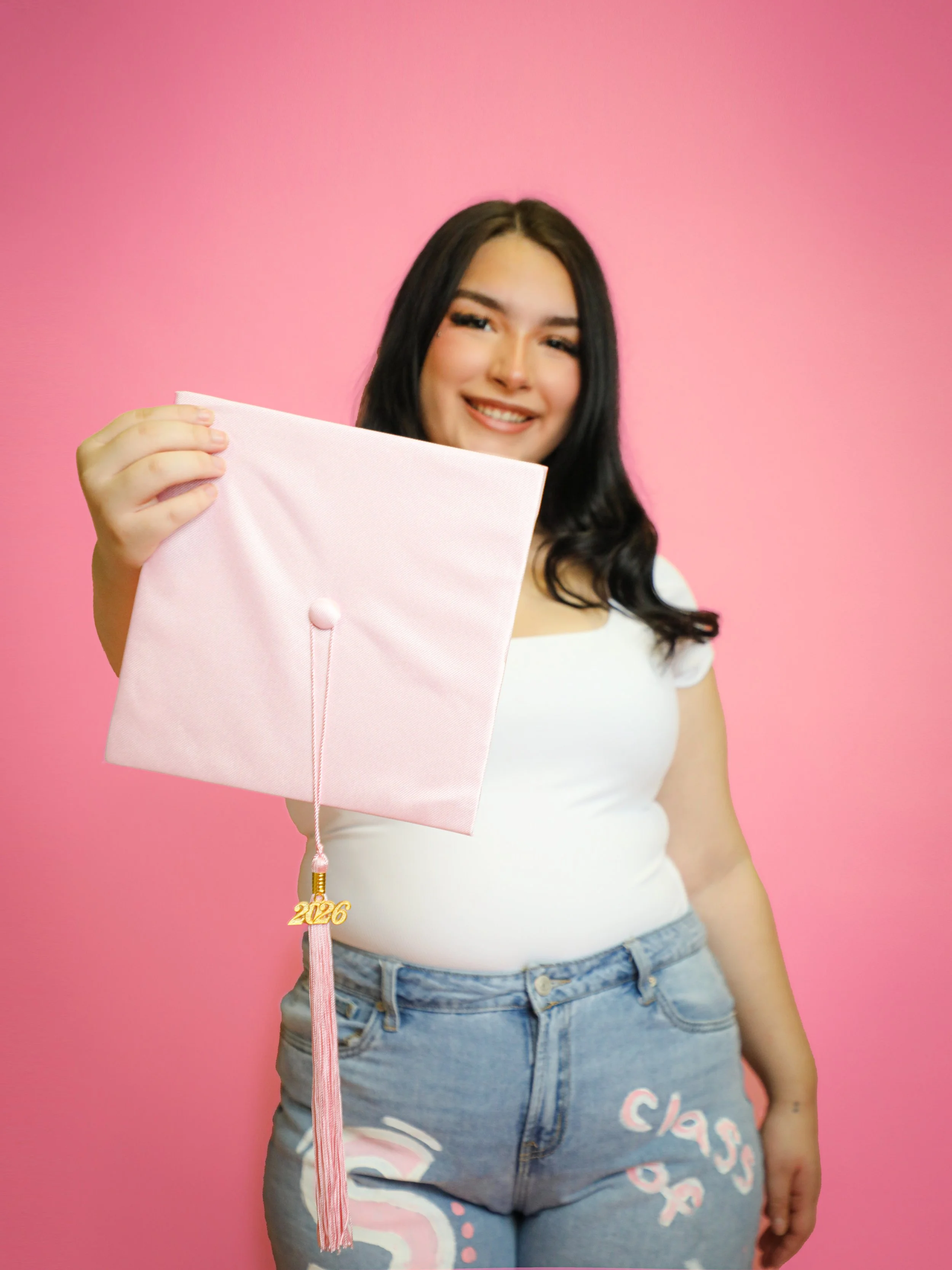 A young woman with long dark hair smiling and holding a pink graduation cap with a "2026" tassel in front of a pink background.