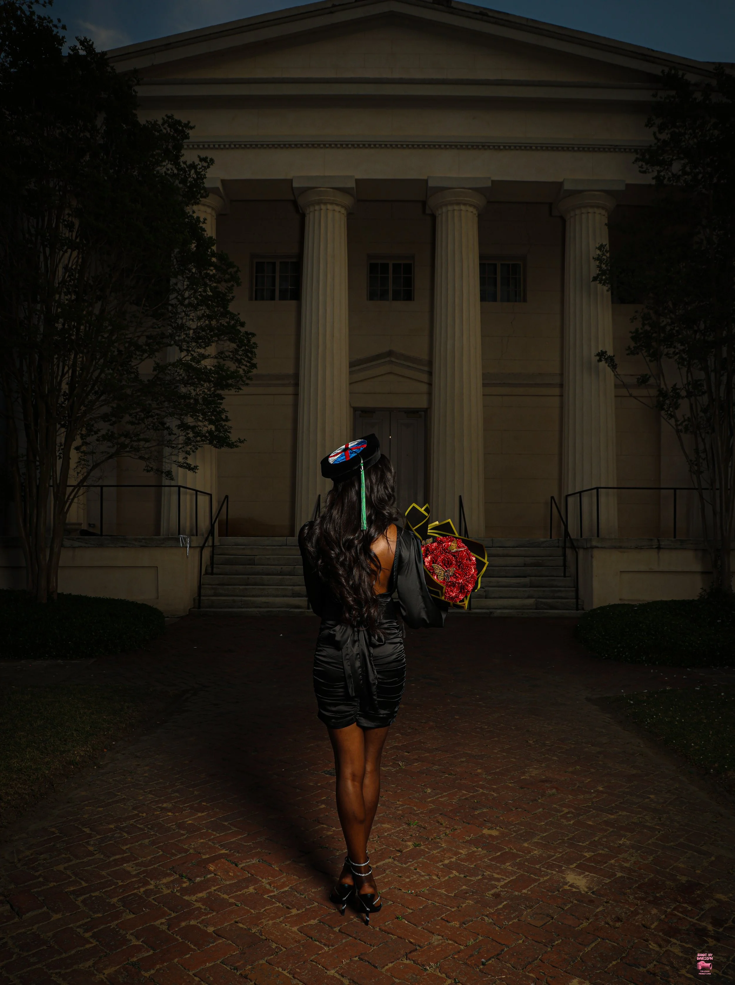 A woman with long dark hair dressed in a black satin dress and high heels, holding a bouquet of flowers, walking towards a historic building with large columns and steps, at night.