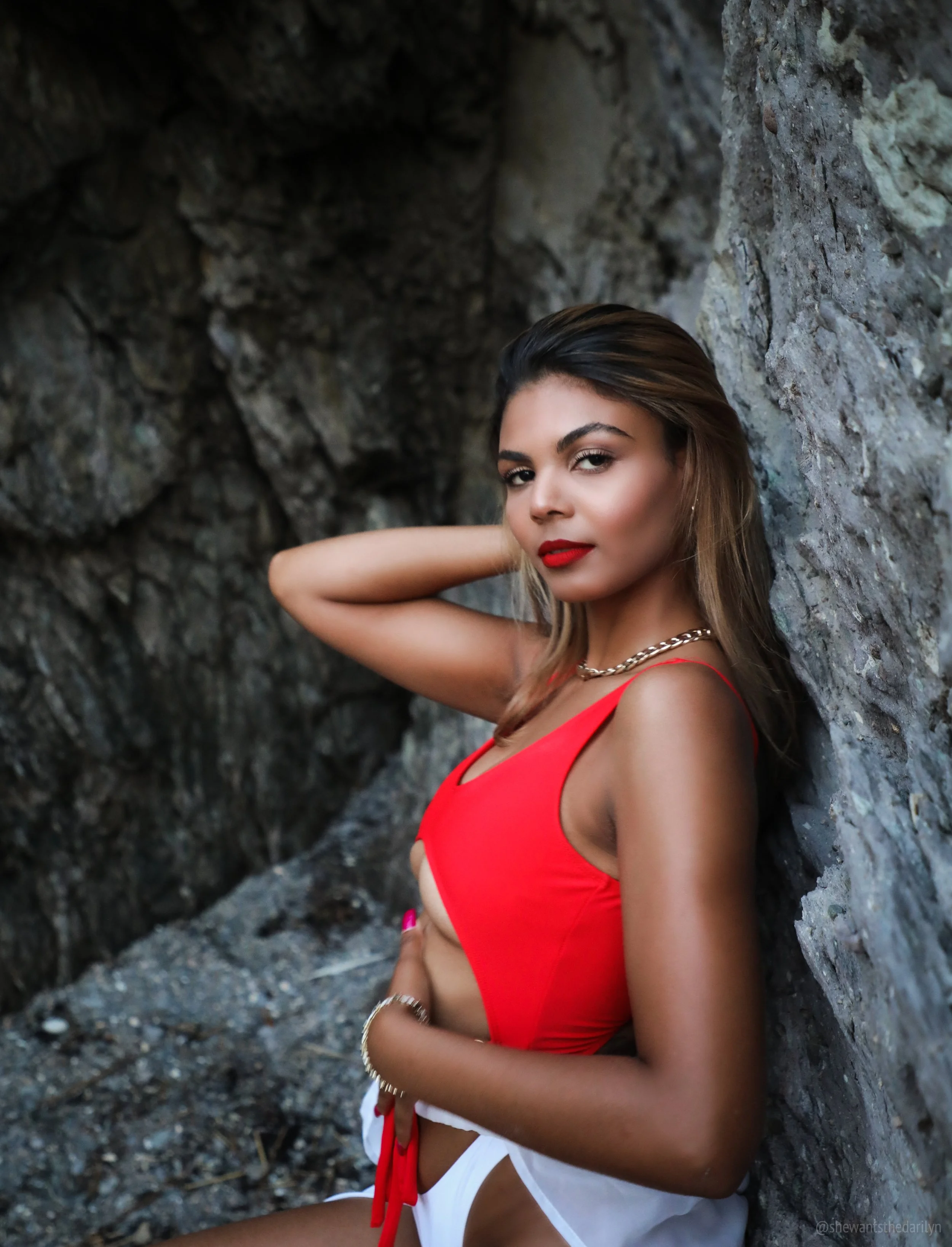 A woman with brown hair, red lipstick, and gold jewelry leaning against a rocky wall, wearing a red tank top and white shorts with a red belt, posing with one hand behind her head and the other on her stomach.