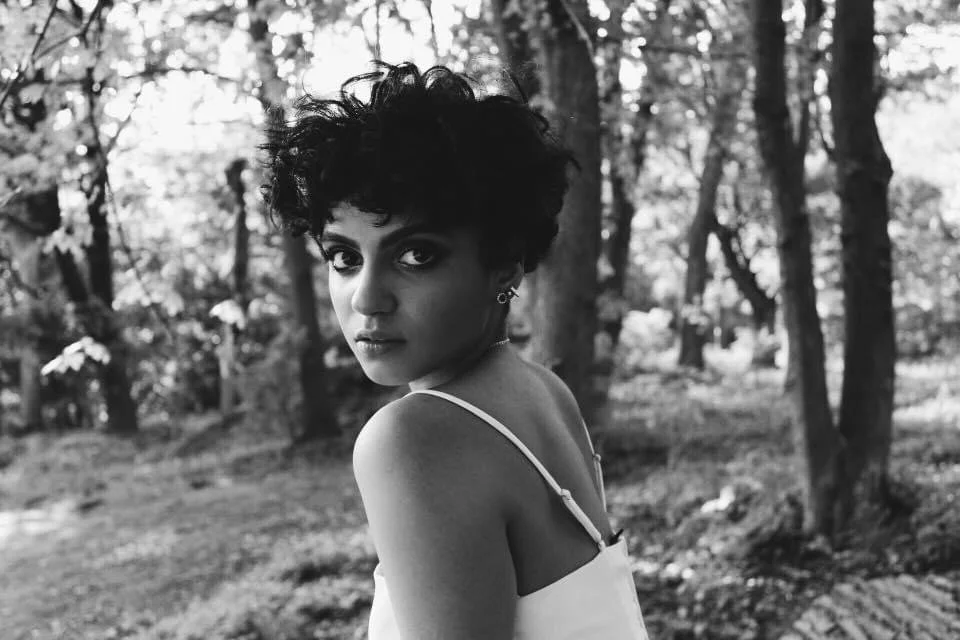 A woman with short, curly hair and earrings looking over her shoulder in a forest setting, black and white photo
