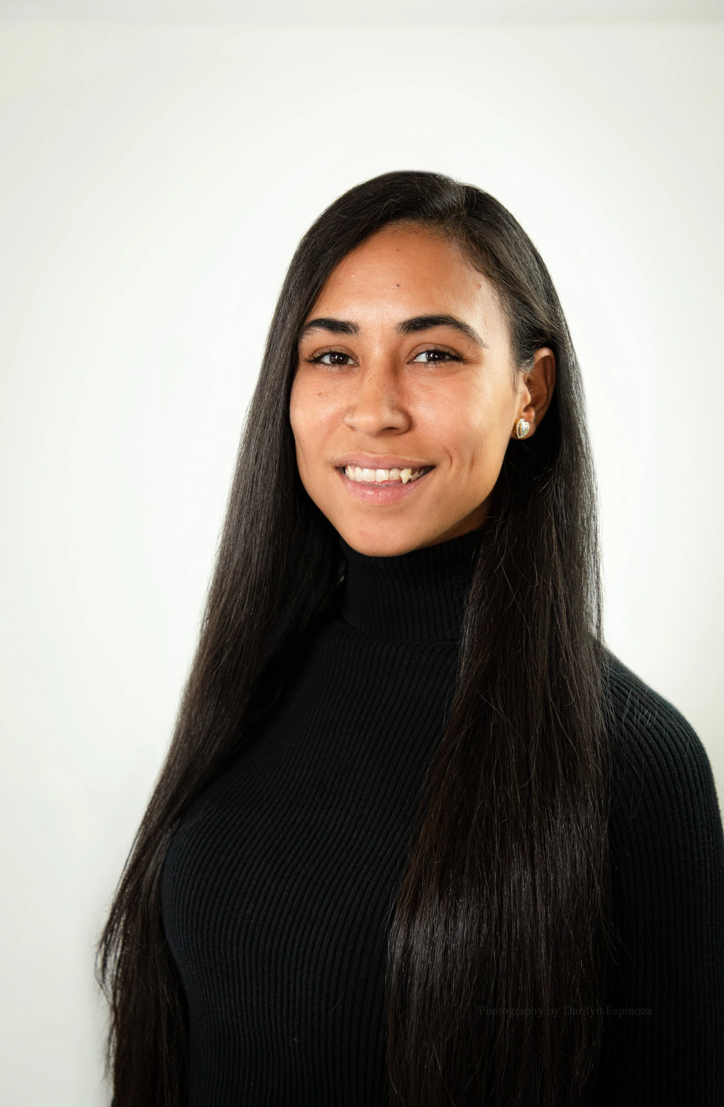 A woman with long dark hair wearing a black turtleneck smiling in front of a plain light background.