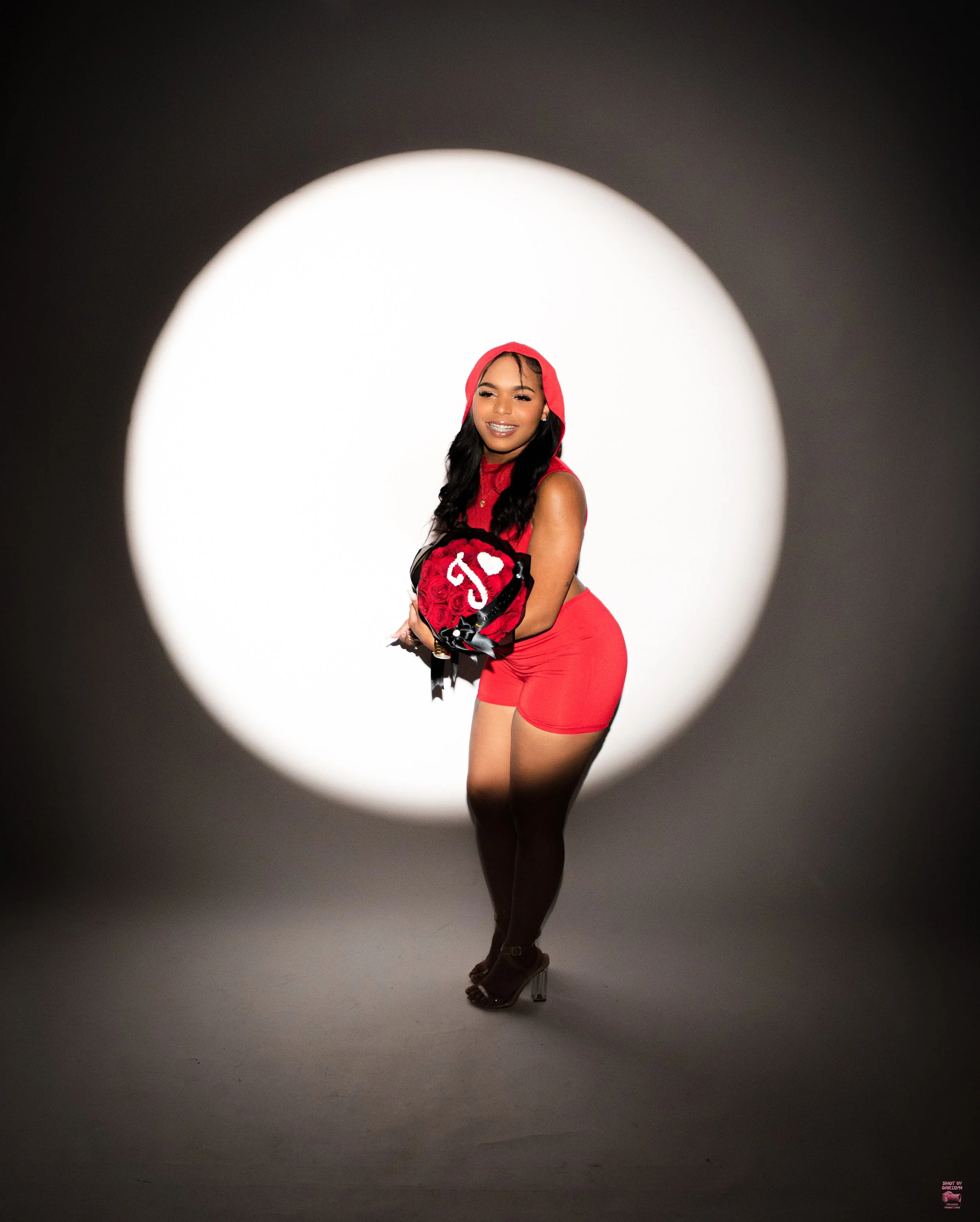 A woman dressed in red shorts and a matching sleeveless hoodie, holding a bouquet of red roses and smiling, captured through a circular cutout against a dark background.