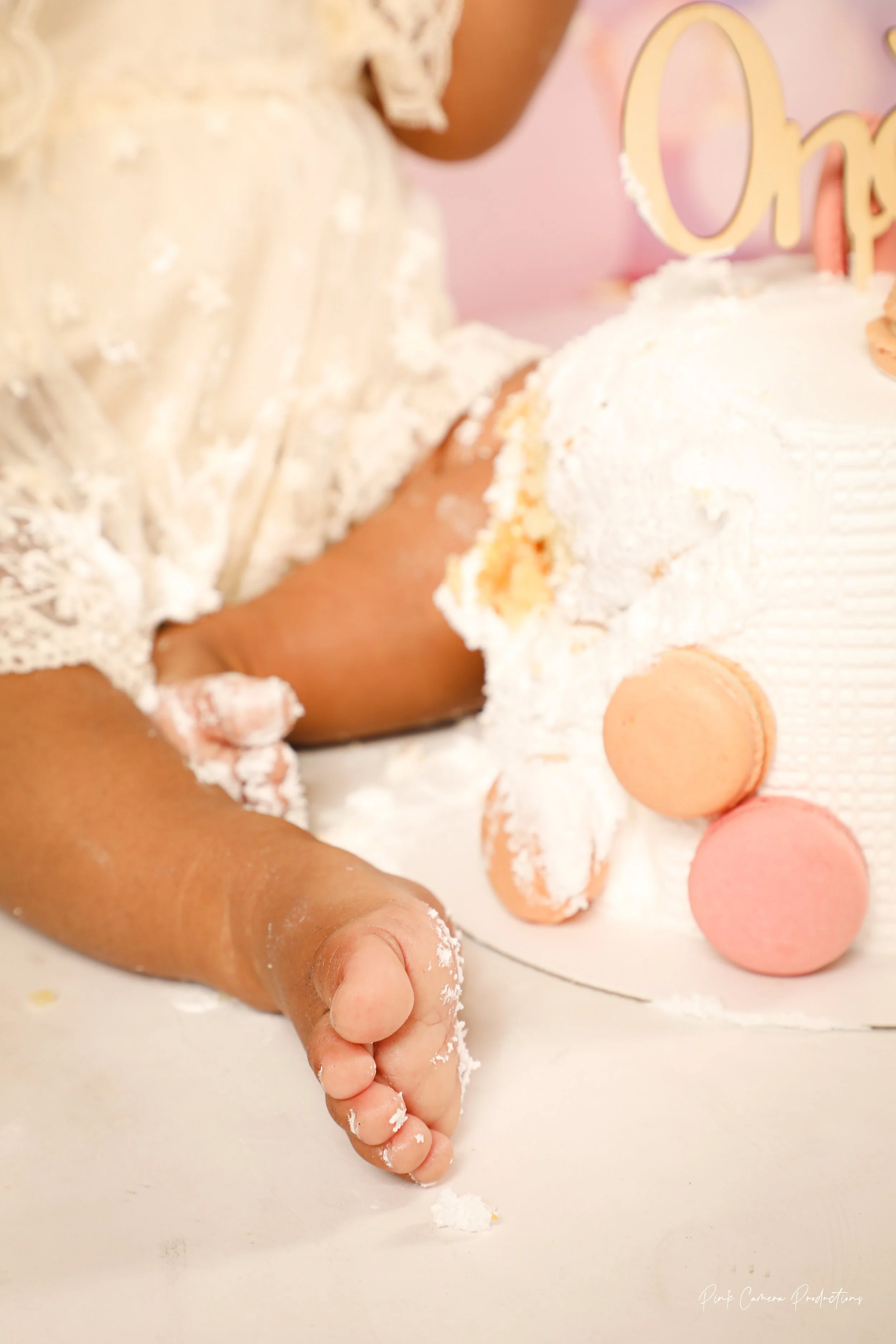 A close-up of a child with her hand covered in white powder, holding a white butter knife and a partially broken cake covered in white frosting with pink and beige macarons on top. The child is wearing a white lace dress, and the background is pink.