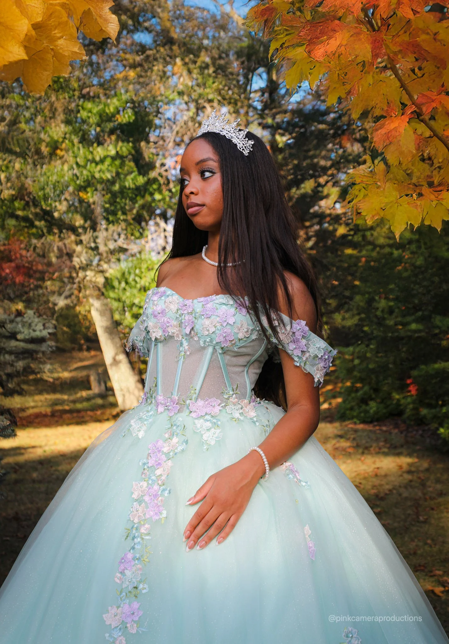 A young woman in an off-shoulder, pastel-colored princess dress with floral embroidery, standing outdoors among autumn trees, wearing a silver tiara, pearl necklace, and bracelet.