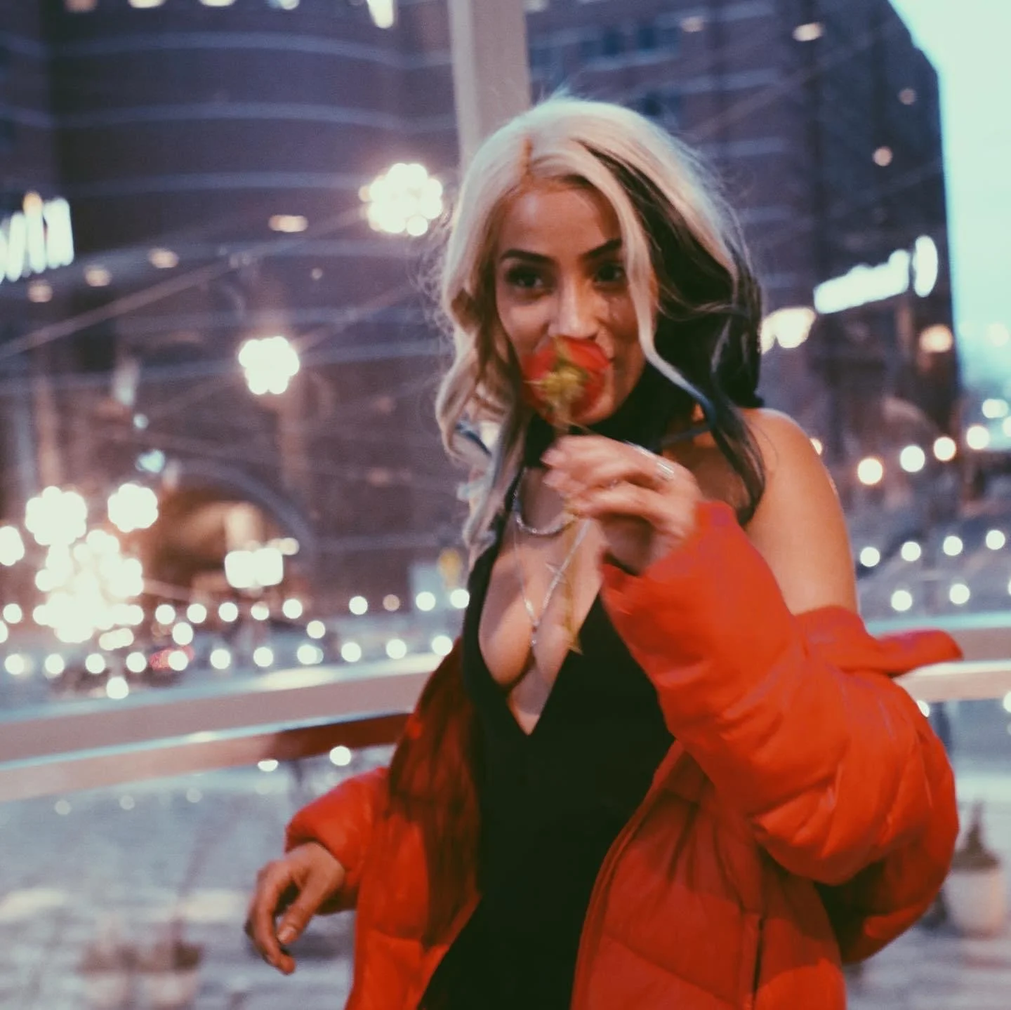 A woman with blonde and black hair eating a strawberry while sitting on a city street at night, with blurred city lights in the background.