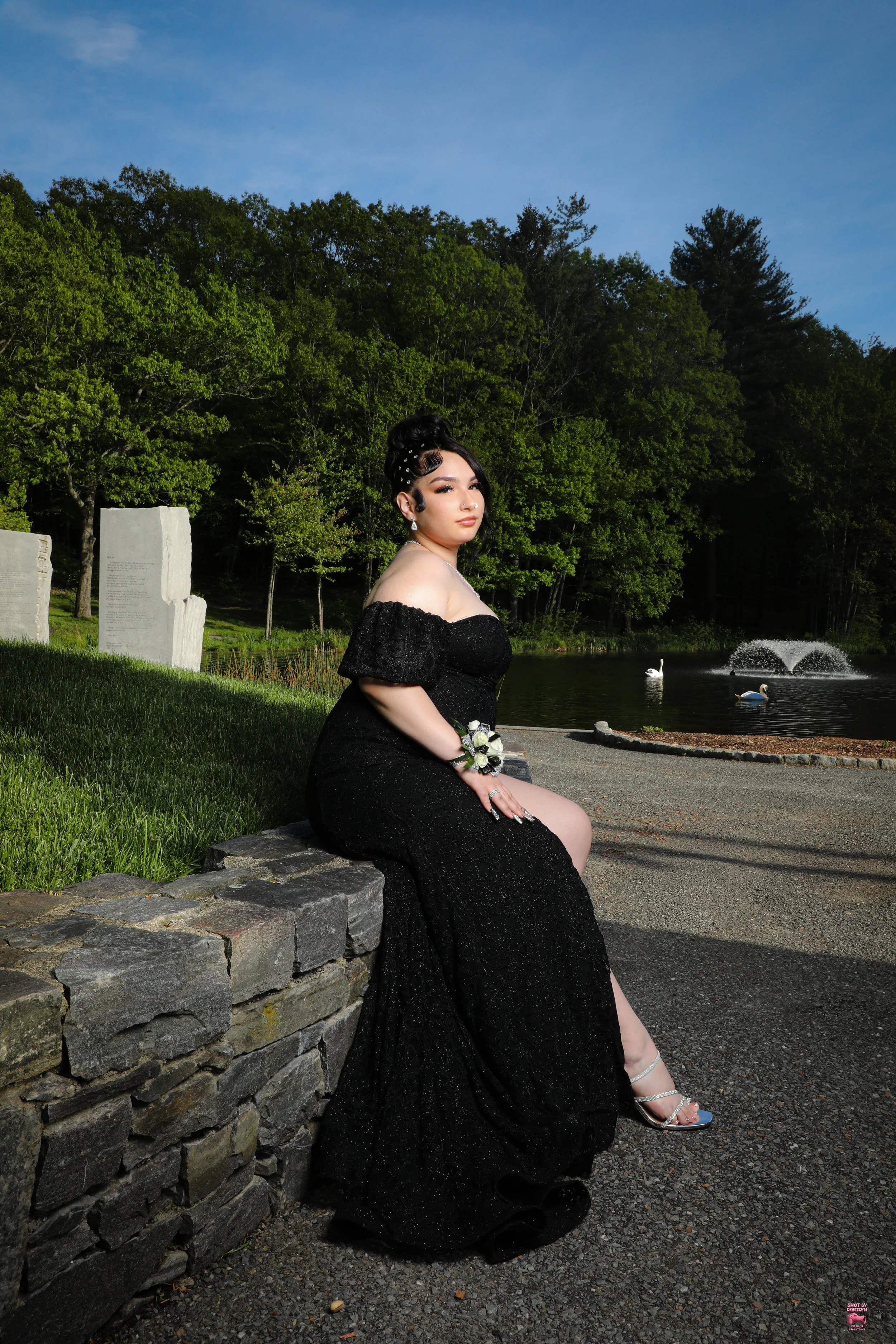 A woman in a black dress sitting on a stone wall by a lake with a fountain and swan-shaped boats, surrounded by green trees under a blue sky.