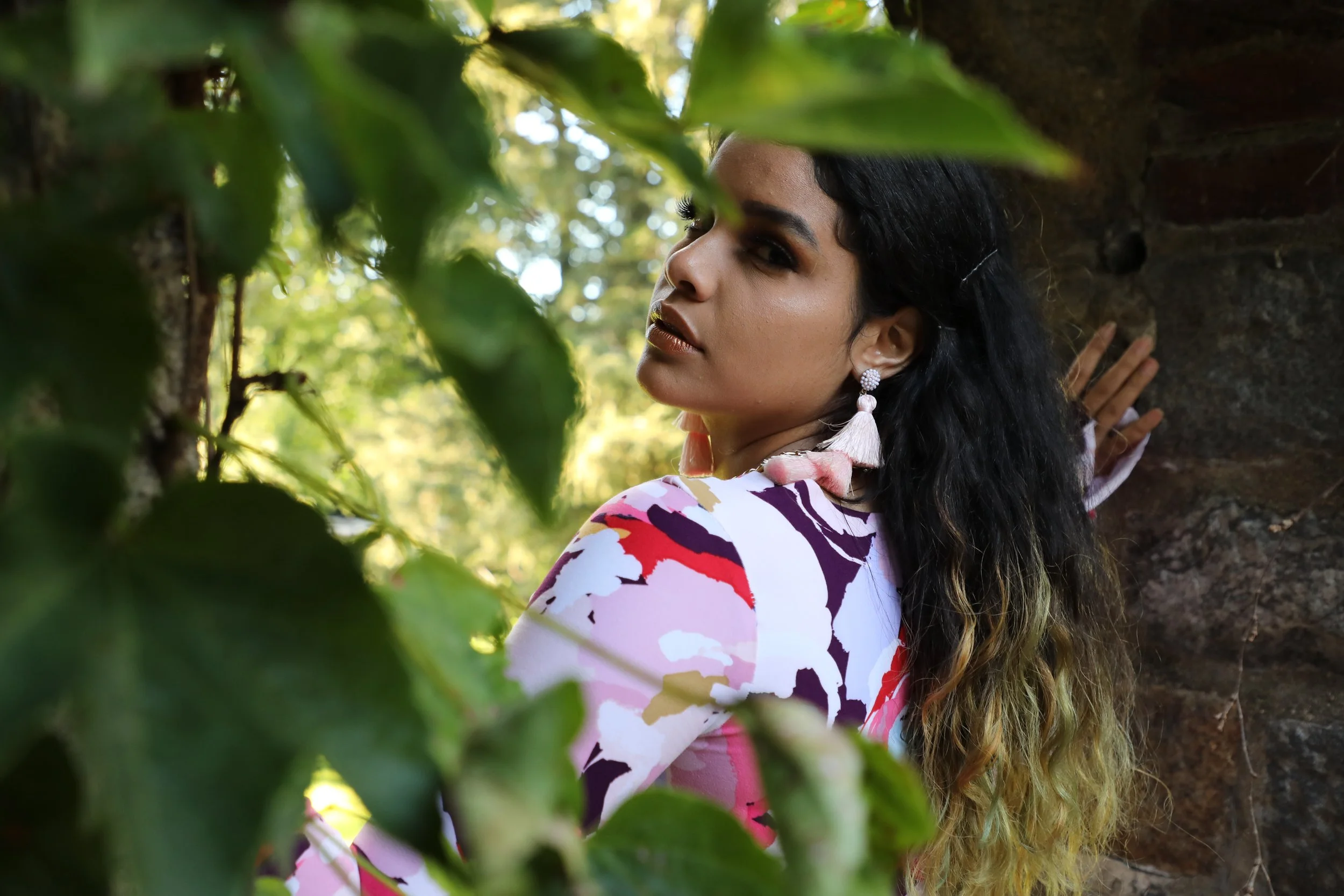 A woman with long curly hair and large earrings, wearing a colorful patterned top, leaning against a rock formation among green foliage, with sunlight filtering through the trees.
