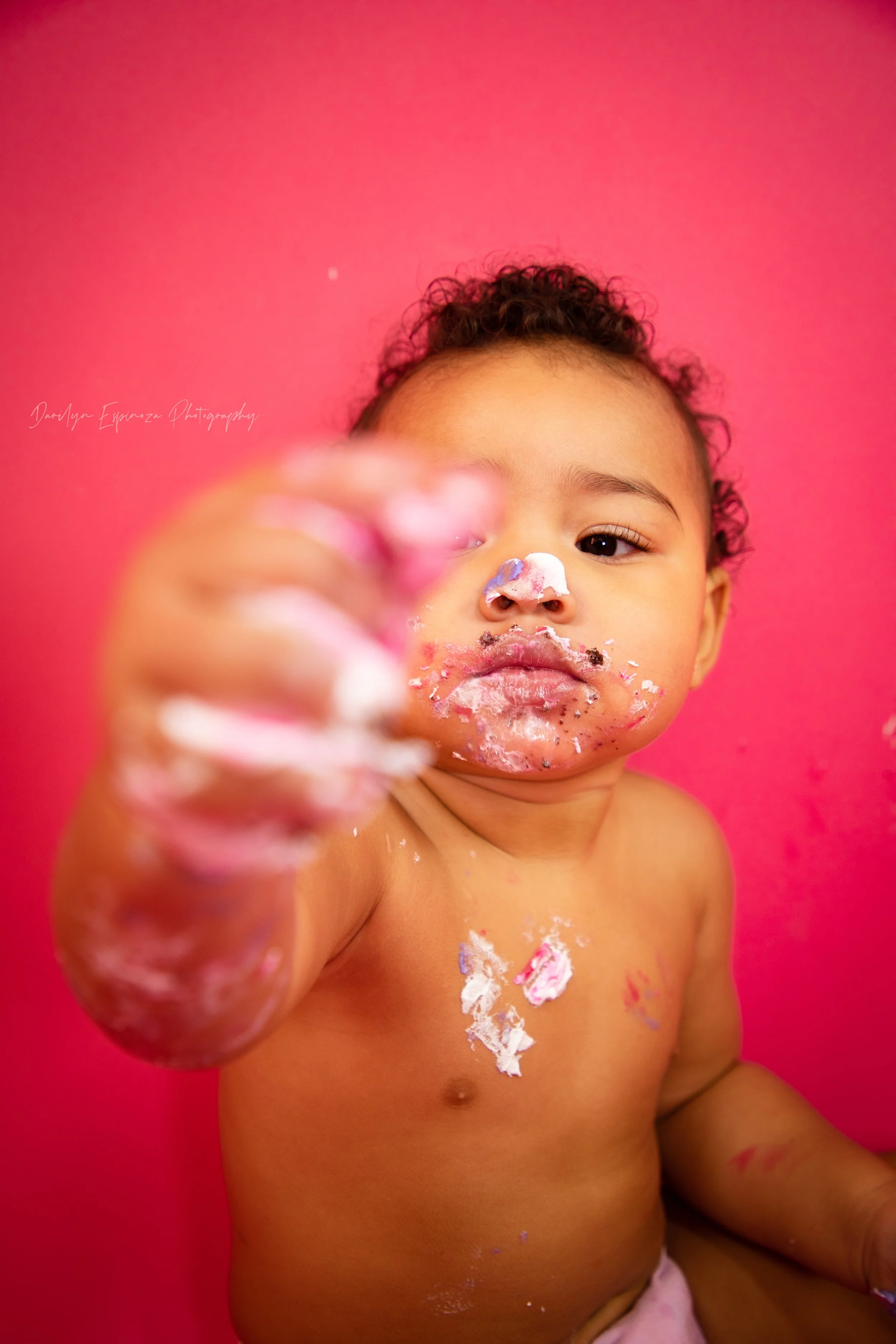 A young child with curly hair and medium skin tone, covered in pink, white, and gray cake or frosting, holding a cake in their hand, against a bright pink background.