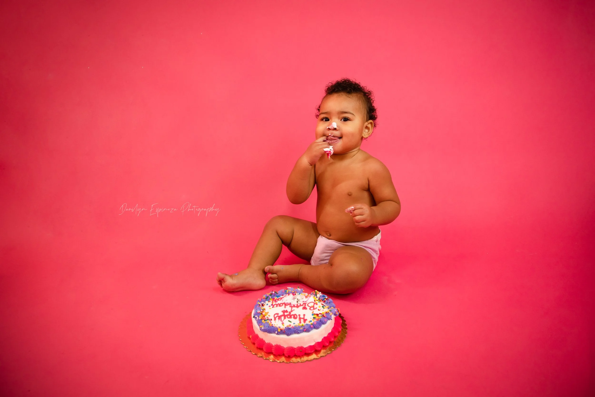 A young toddler sitting on a pink background next to a decorated birthday cake, with frosting on their face and wearing only a diaper.