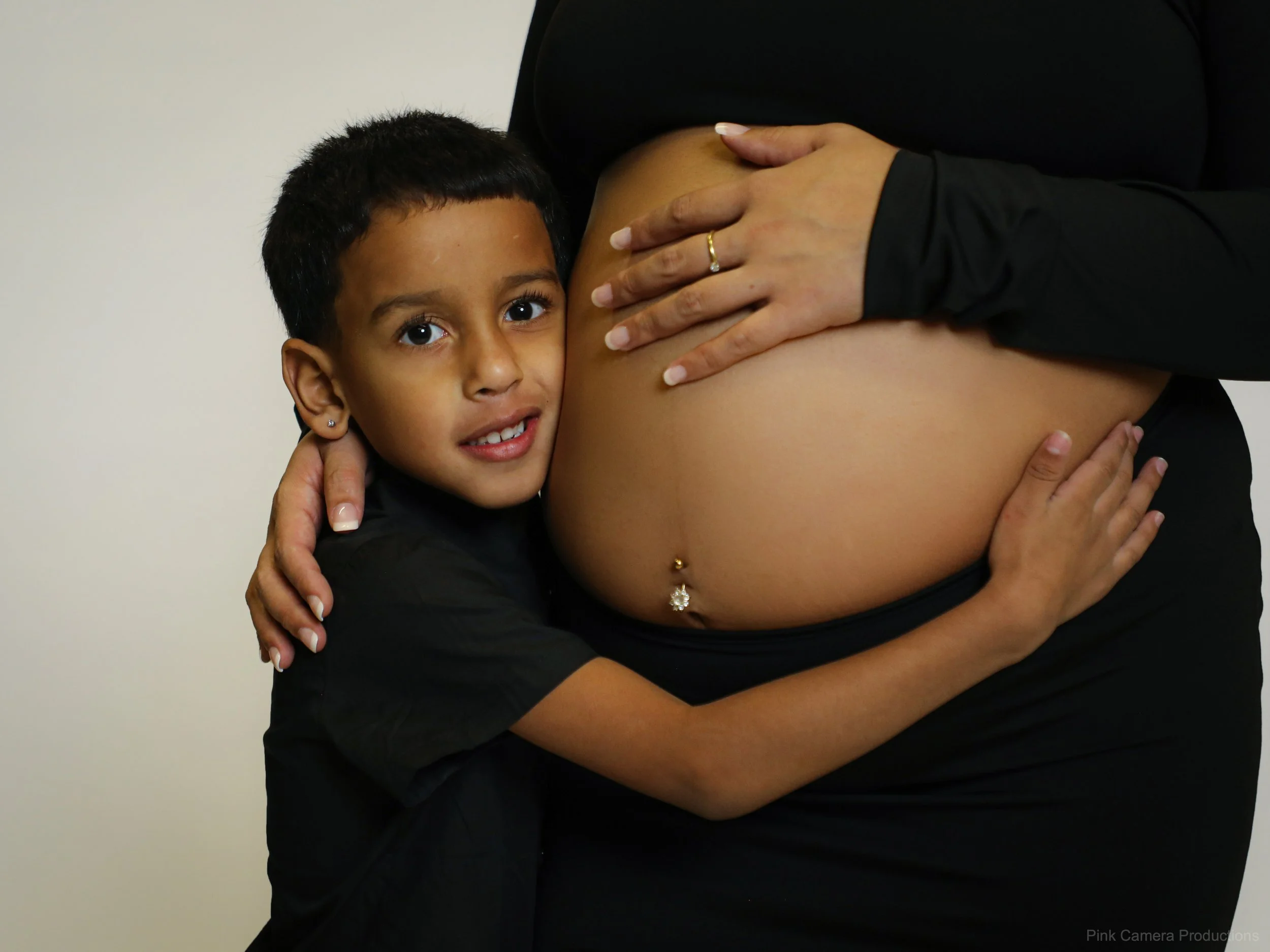 A young boy hugging a pregnant woman's belly, with her hand resting on her stomach, in a black long-sleeve shirt. The boy has short curly hair, earrings, and is looking at the camera.