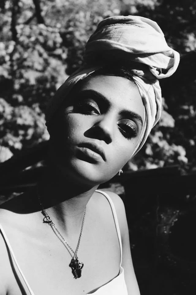 Black and white photo of a woman wearing a headscarf, necklace, and a sleeveless top, with sunlight casting shadows on her face outdoors.