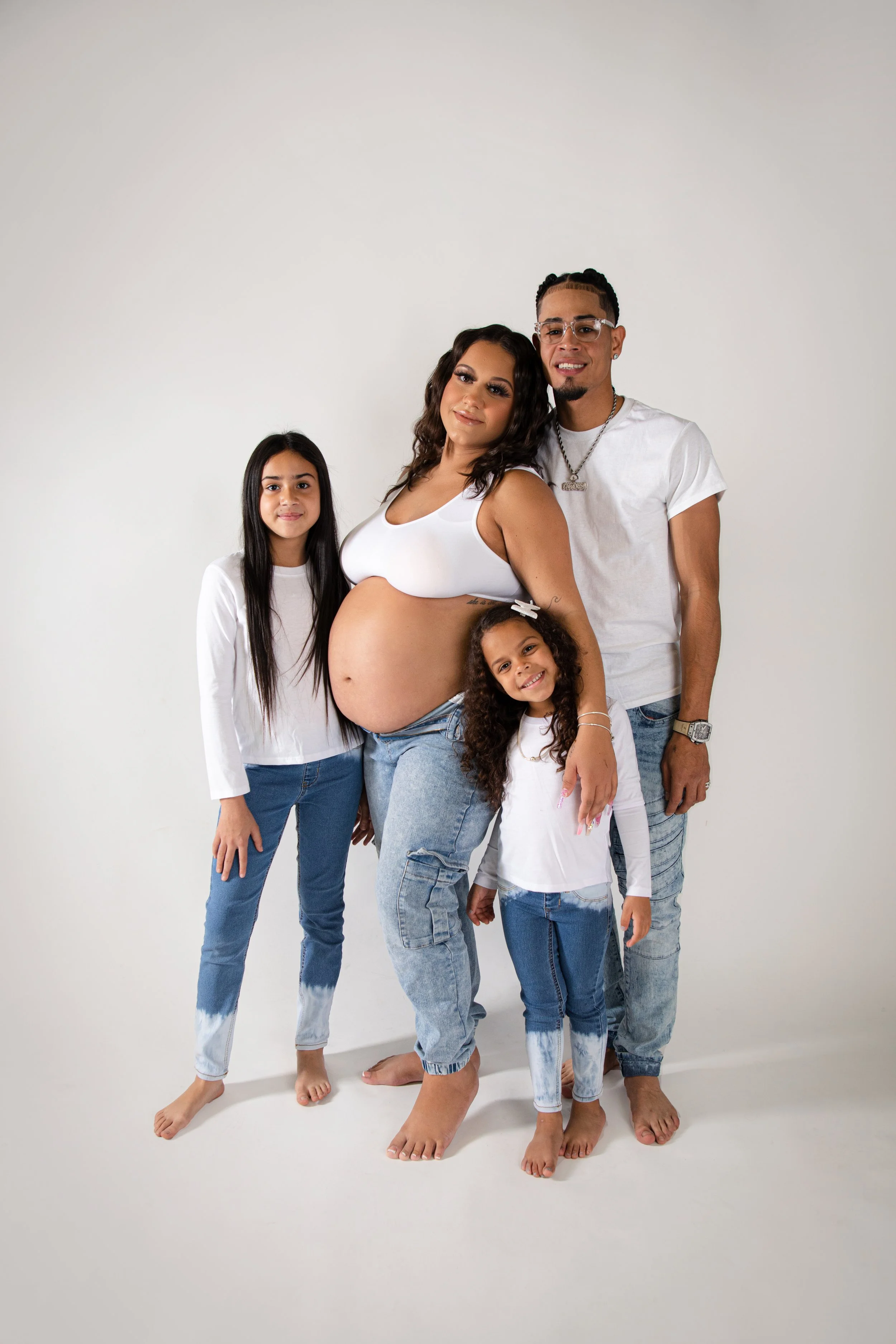 Family of five, including pregnant woman, standing together against a plain white background, all dressed in white and blue, barefoot, smiling at the camera.