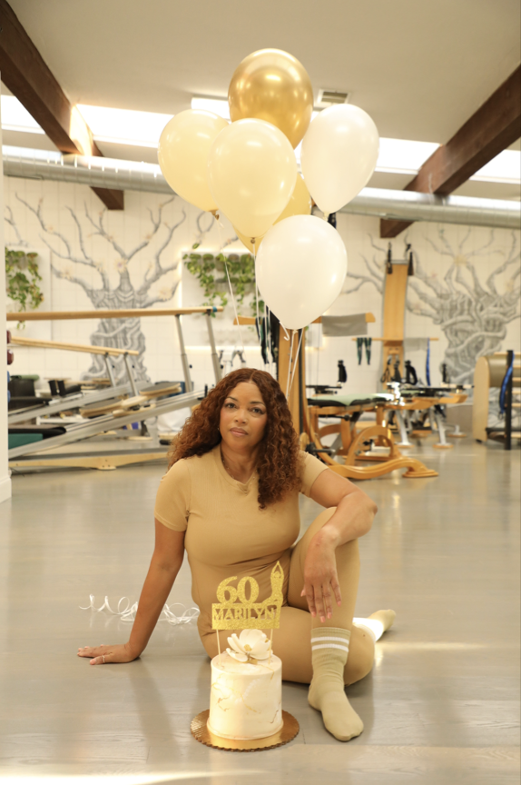 A woman with curly hair sitting on the floor of a Pilates studio, celebrating her 60th birthday with a cake, balloons, and a "60 Marilyn" decoration.