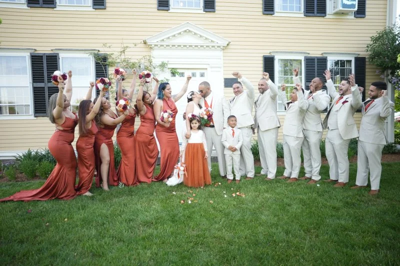 Group of wedding party members including bridesmaids in rust-colored dresses and groomsmen in white suits celebrating outdoors in front of a house.