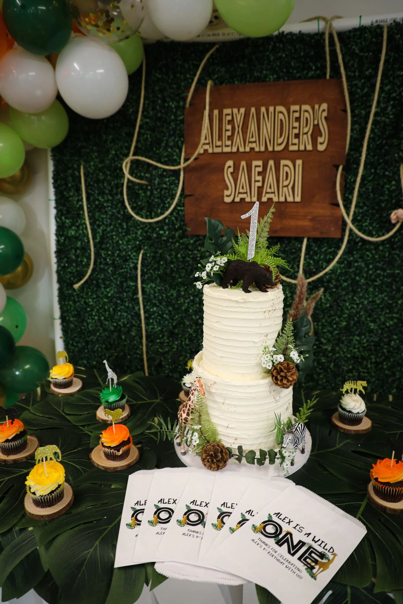 A jungle-themed birthday party setup with a tall, white, textured cake decorated with dinosaur and animal figurines, pinecones, and greenery. The backdrop features a wooden sign reading 'Alexander's Safari' and a balloon arch in shades of green, whit