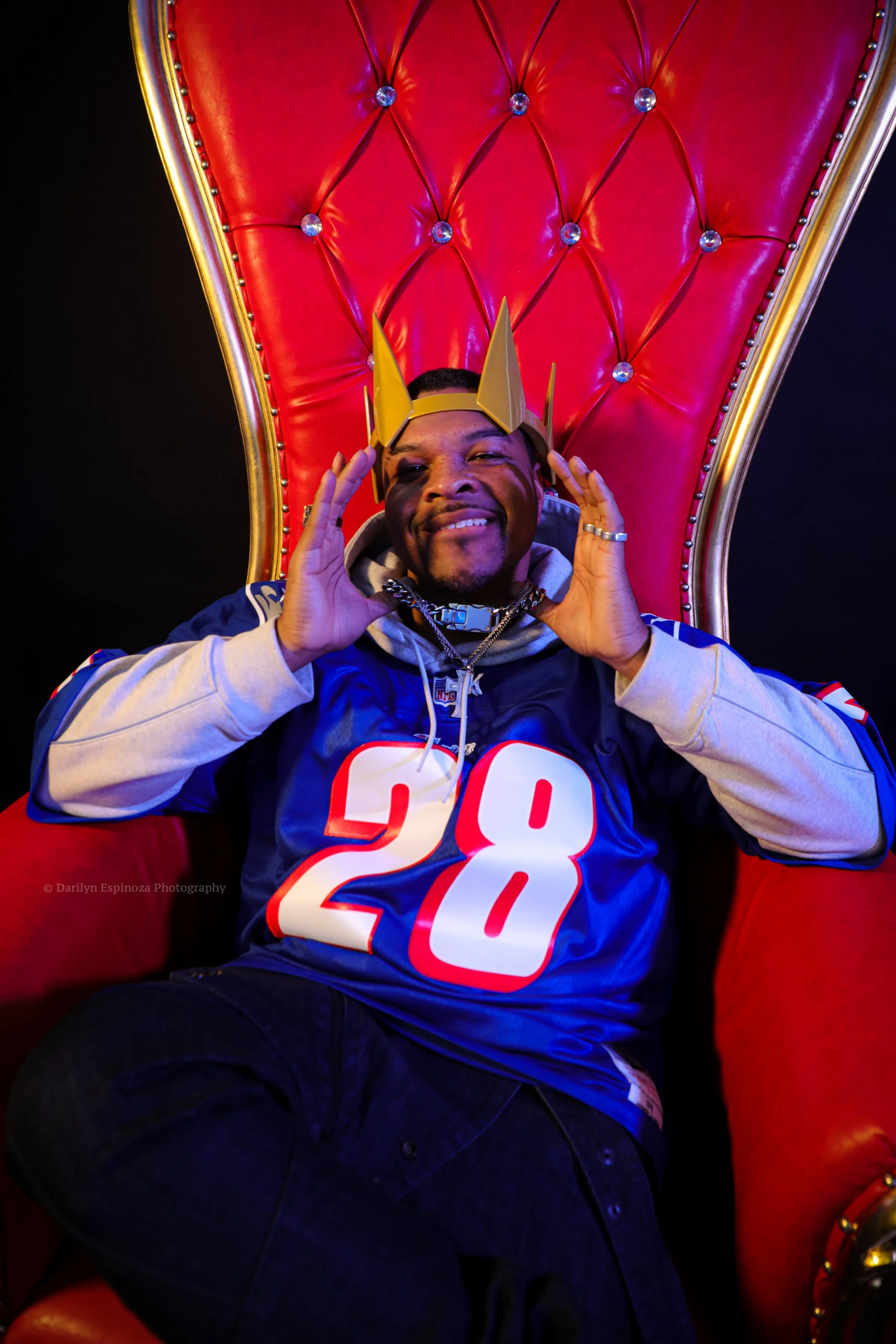 Man wearing a Philadelphia Eagles football jersey sitting on a red throne-like chair, wearing a yellow crown, smiling and holding his hands near his head.