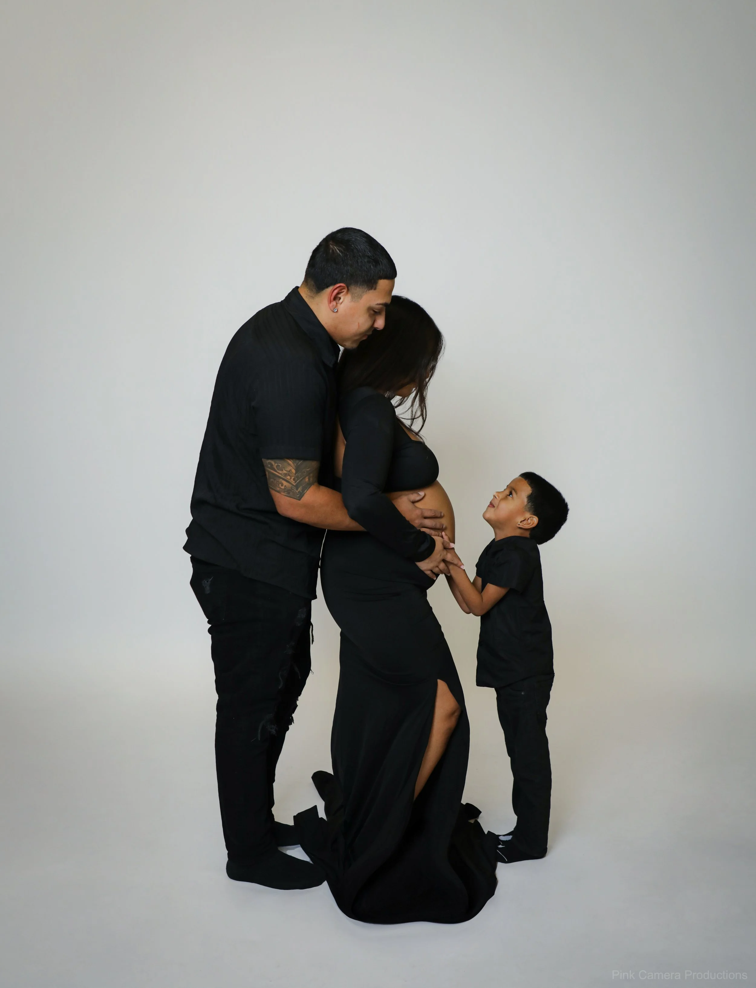 A pregnant woman dressed in a black gown with a slit, kneeling, is being embraced by a man behind her. A young boy standing in front of her holds her hand, looking up at her affectionately. All are dressed in black against a plain light background.
