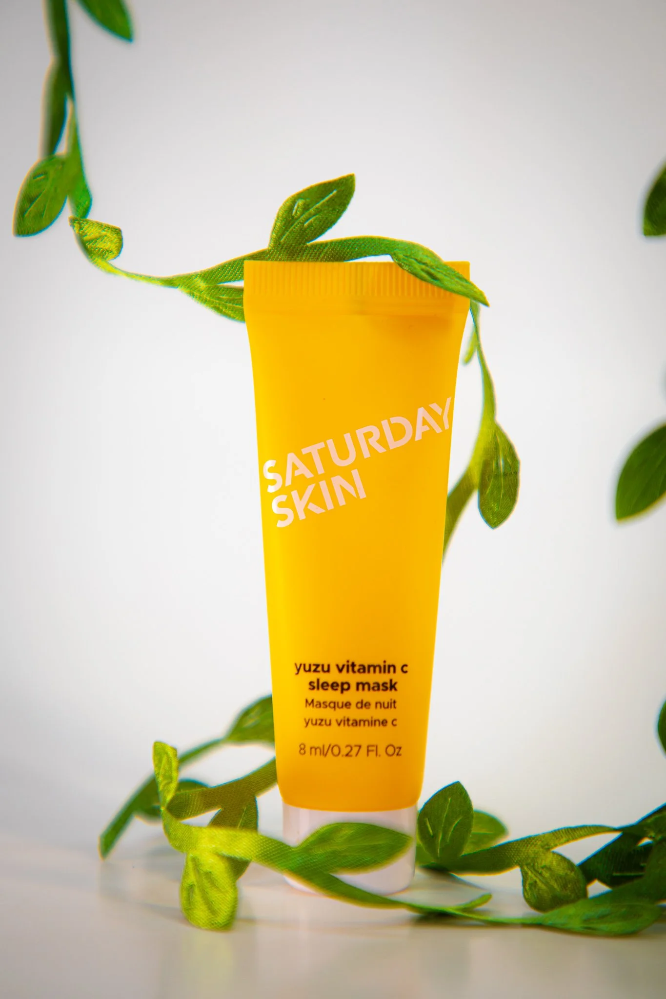 A yellow tube of Saturday Skin yuzu vitamin C sleep mask surrounded by green plant leaves.