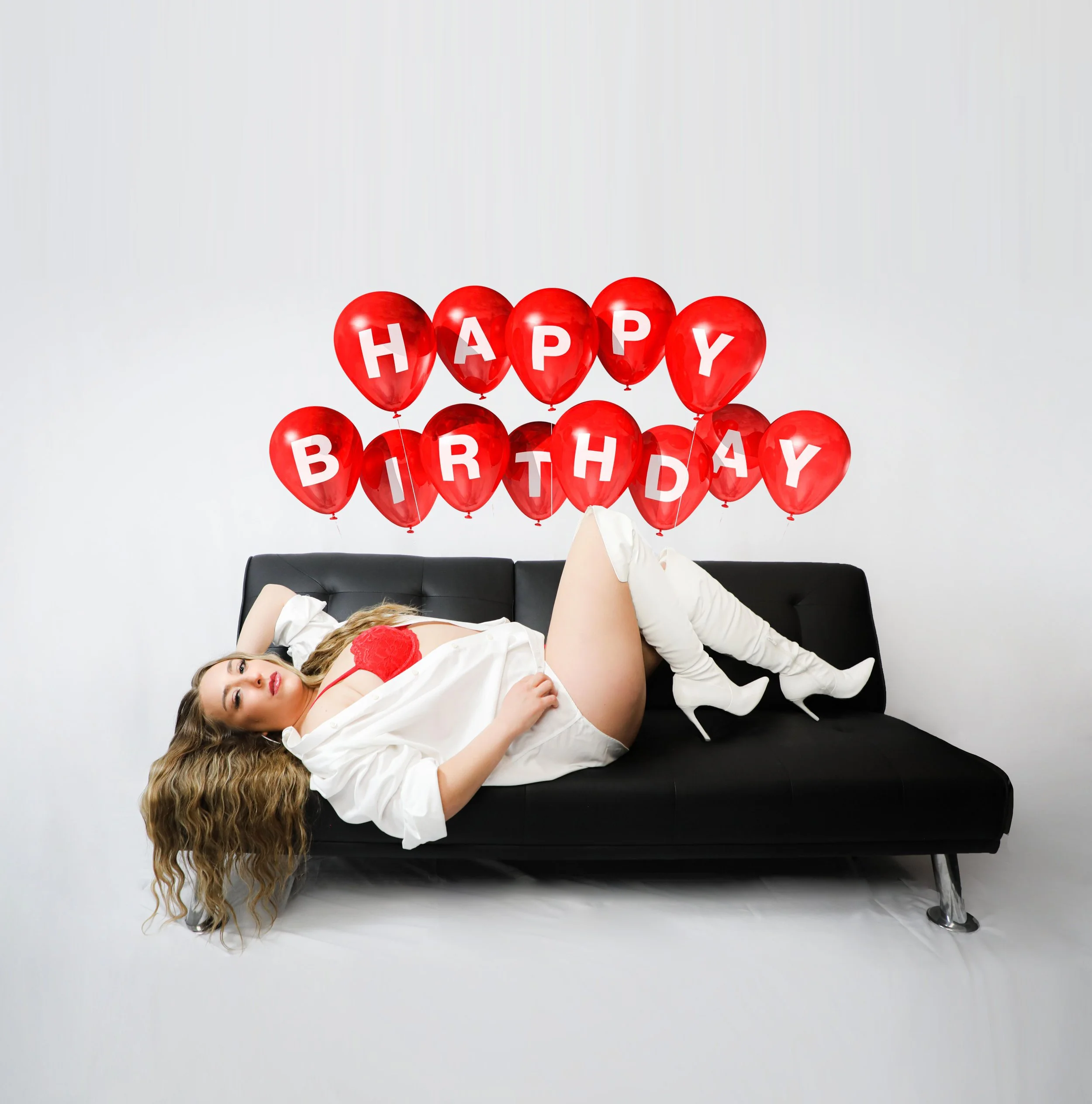 A woman with long wavy hair lying on a black sofa, wearing white thigh-high boots and a white outfit with red lingerie detail, surrounded by red balloons spelling 'HAPPY BIRTHDAY'. the background is plain white.