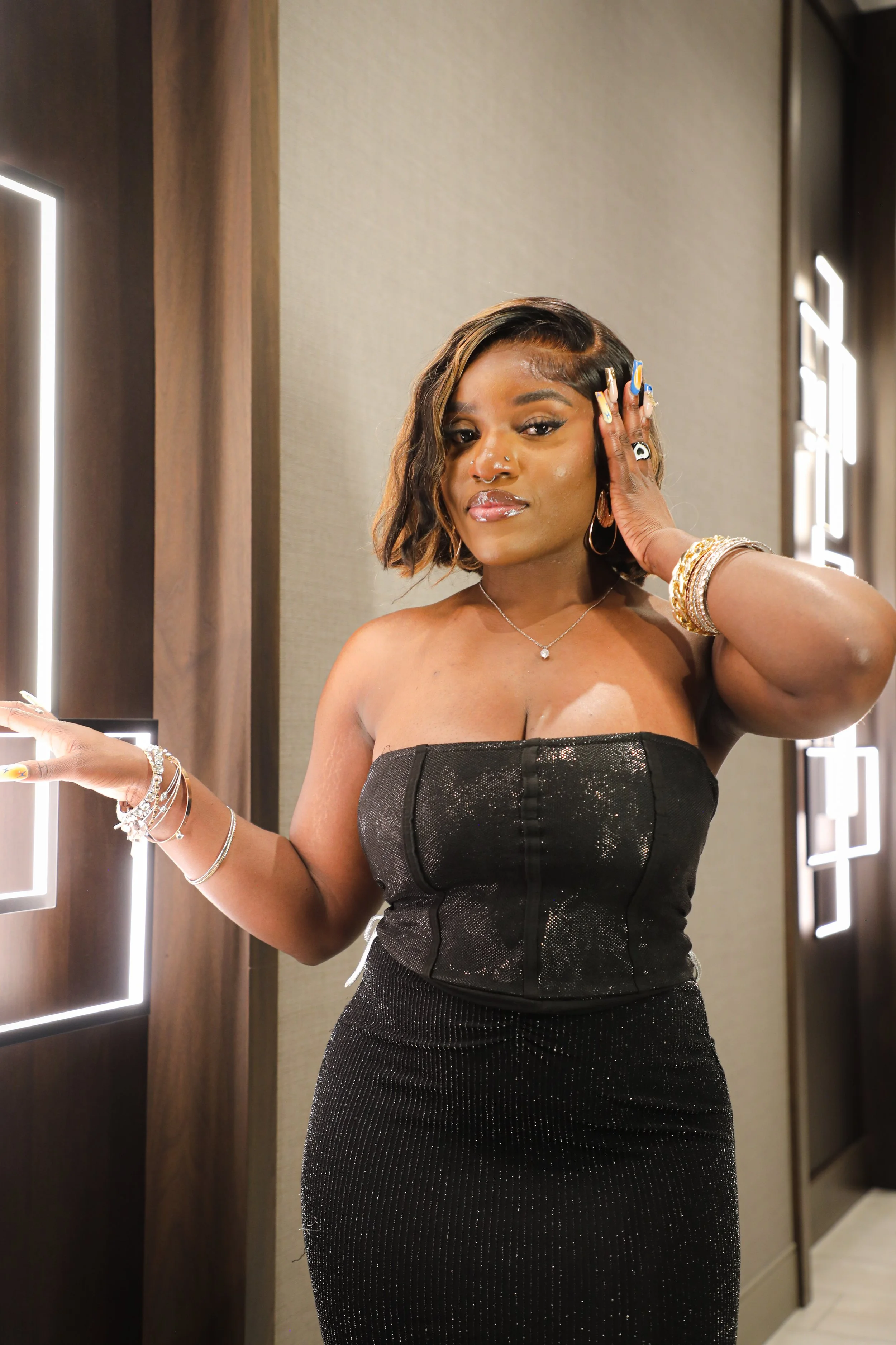 A woman in a black strapless dress standing in front of illuminated wall decor, touching her face with her right hand, wearing jewelry and makeup.