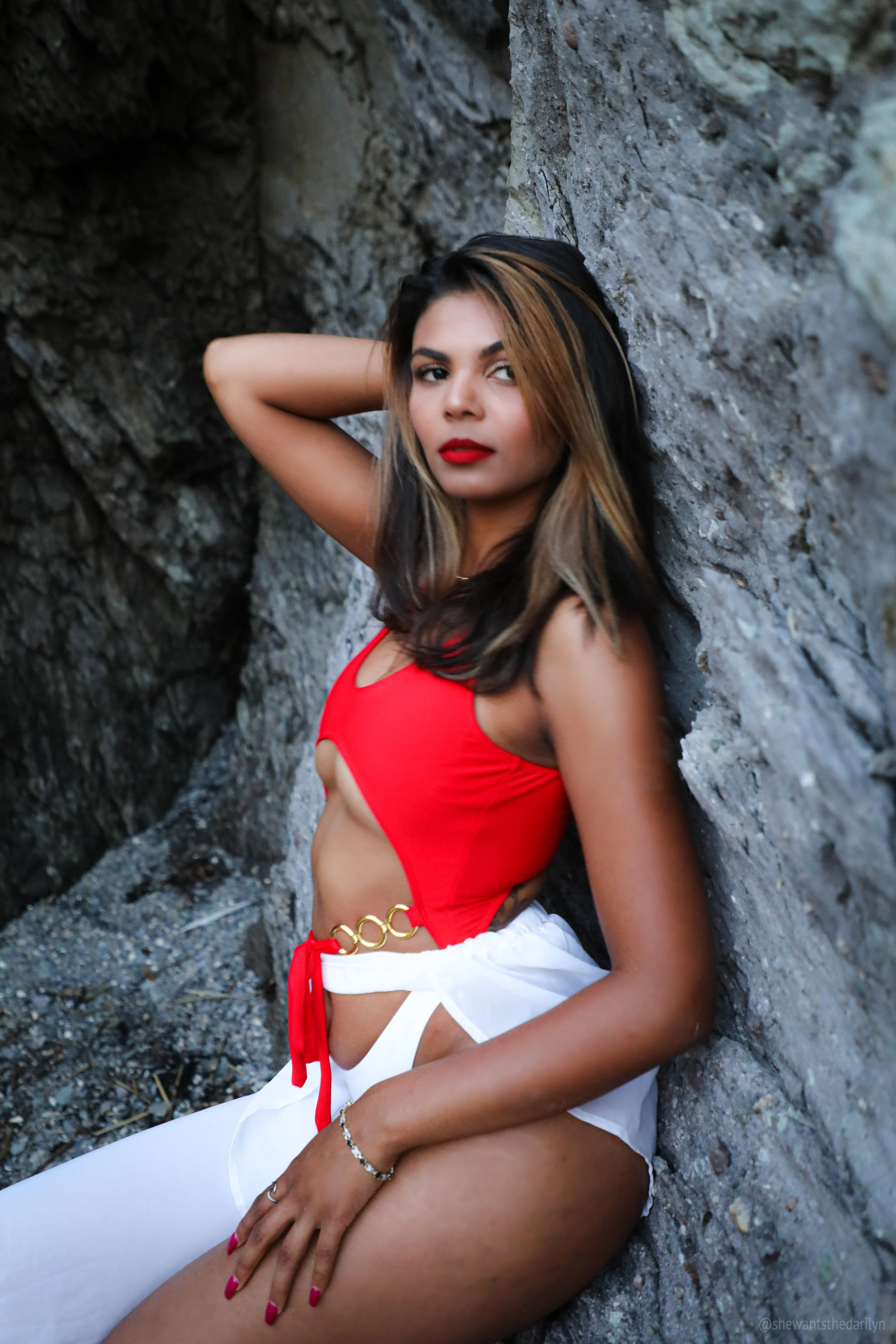 A woman with wavy hair and red lipstick, wearing a red sleeveless top and white shorts, sitting against a rocky wall with one arm behind her head.