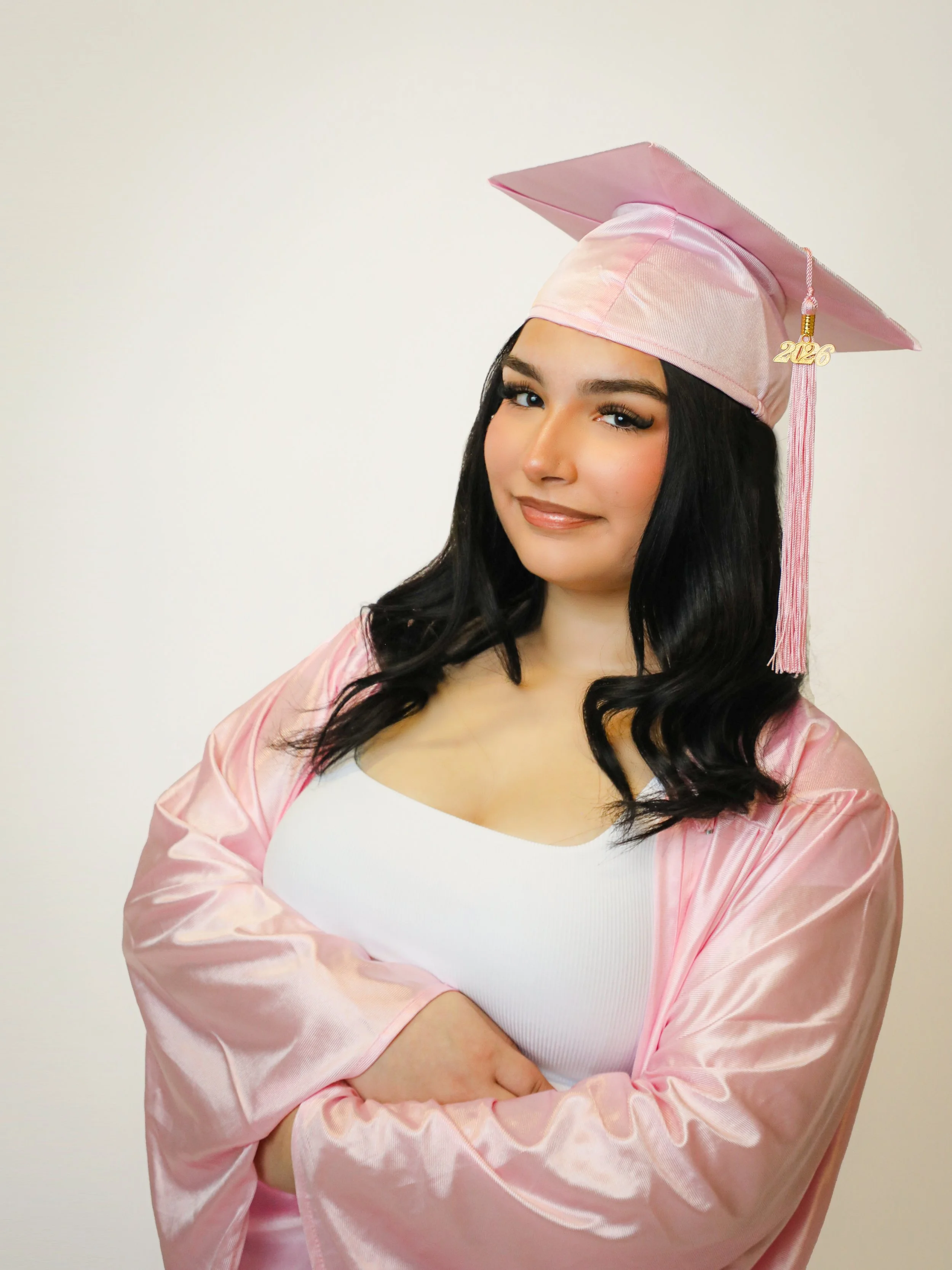 A young woman wearing a pink graduation cap and gown, smiling with her arms crossed, standing against a plain white background.