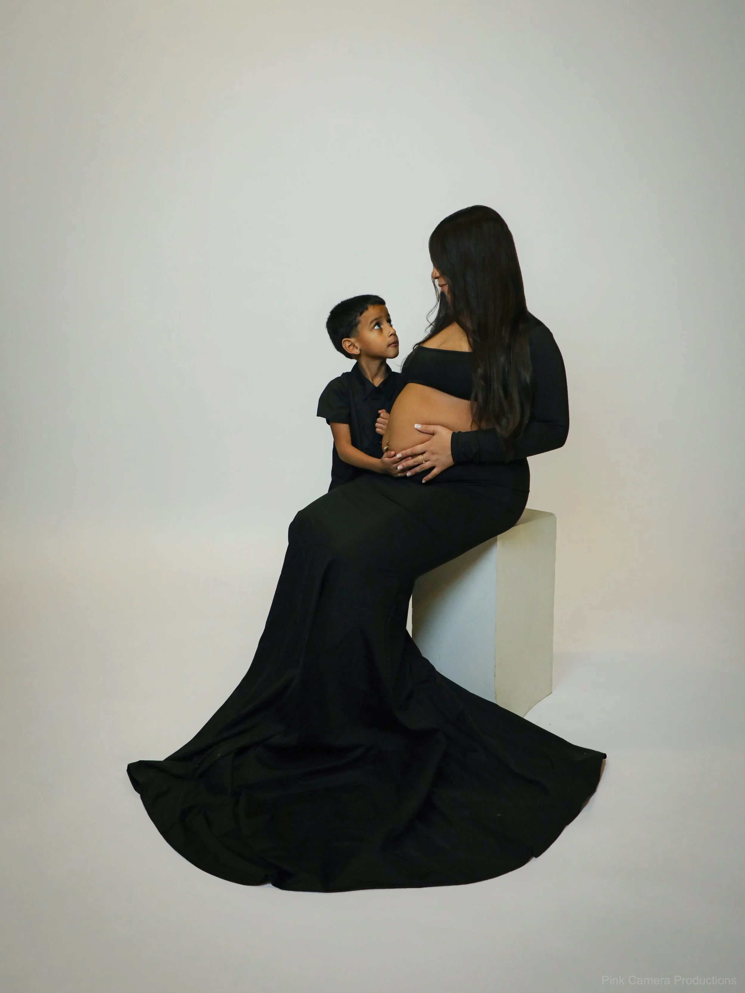 A pregnant woman with long dark hair and a young boy sitting on a white pedestal against a plain white background, looking at each other.