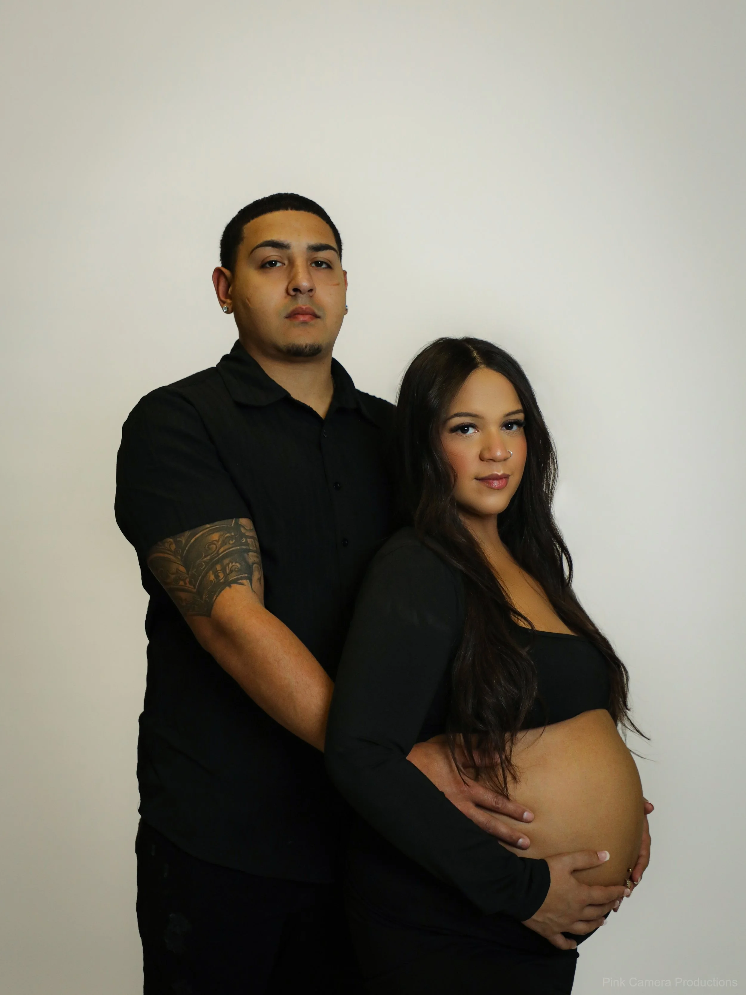 A couple poses in front of a plain white wall; she is pregnant, wearing a black top with a smile, and he is in a black shirt with a serious expression, standing behind her with his hands on her belly.