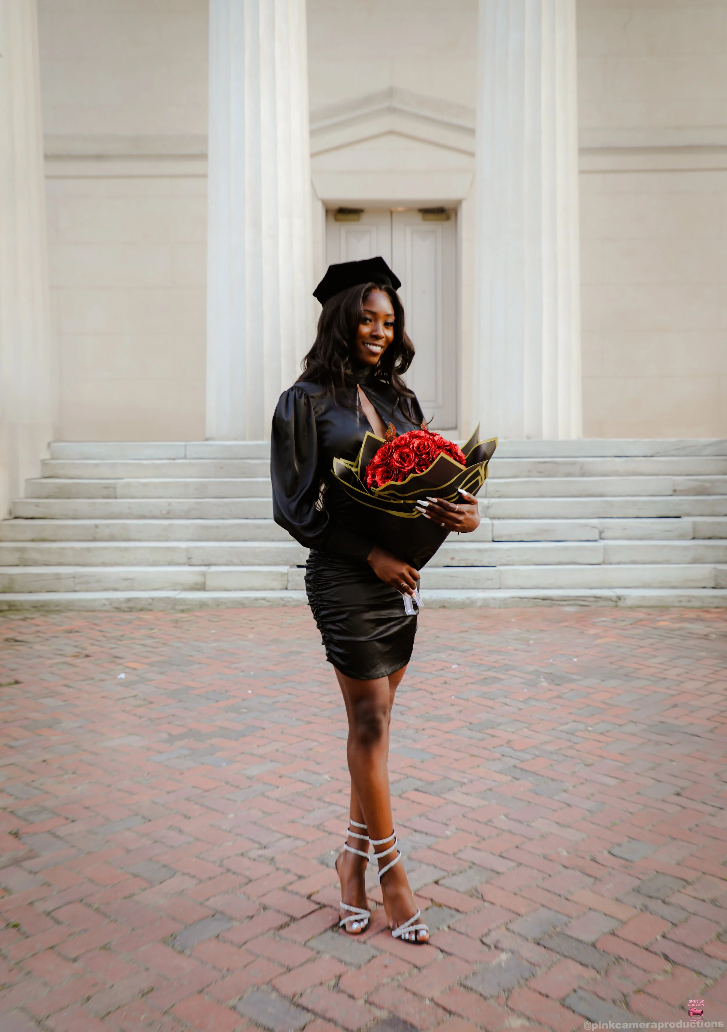 A young woman in a graduation cap and gown holding a bouquet of red roses, standing on brick steps in front of a classical building with pillars.