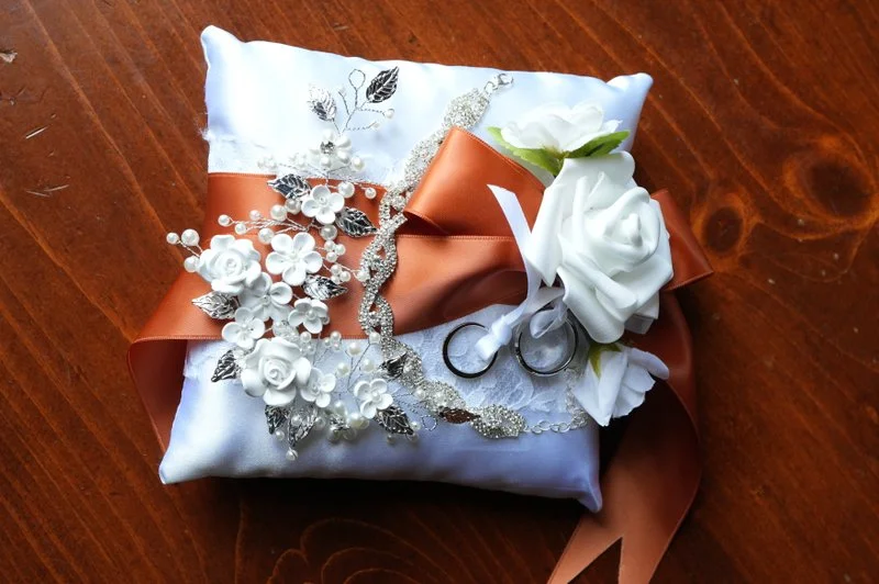 Gift wrapped in white satin with white flowers, orange ribbon, silver jewelry, and a pair of small scissors, placed on a wooden surface.