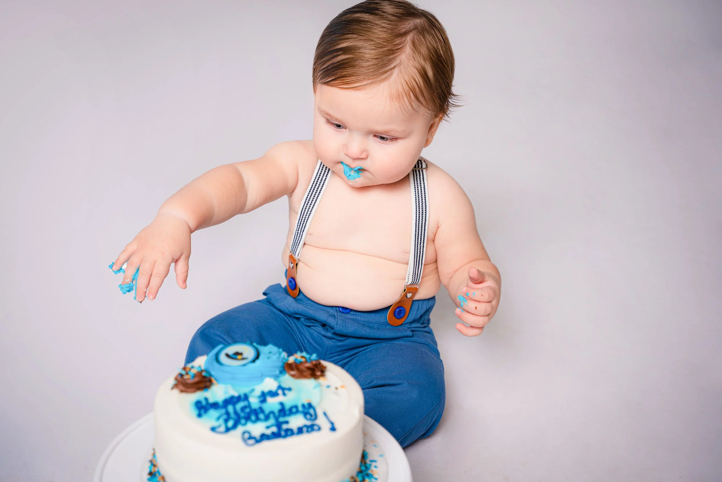 A young toddler wearing blue pants and suspenders, sitting on the floor, with blue cake smudged on his face and hands in front of a birthday cake with blue decorations, on a plain gray background.