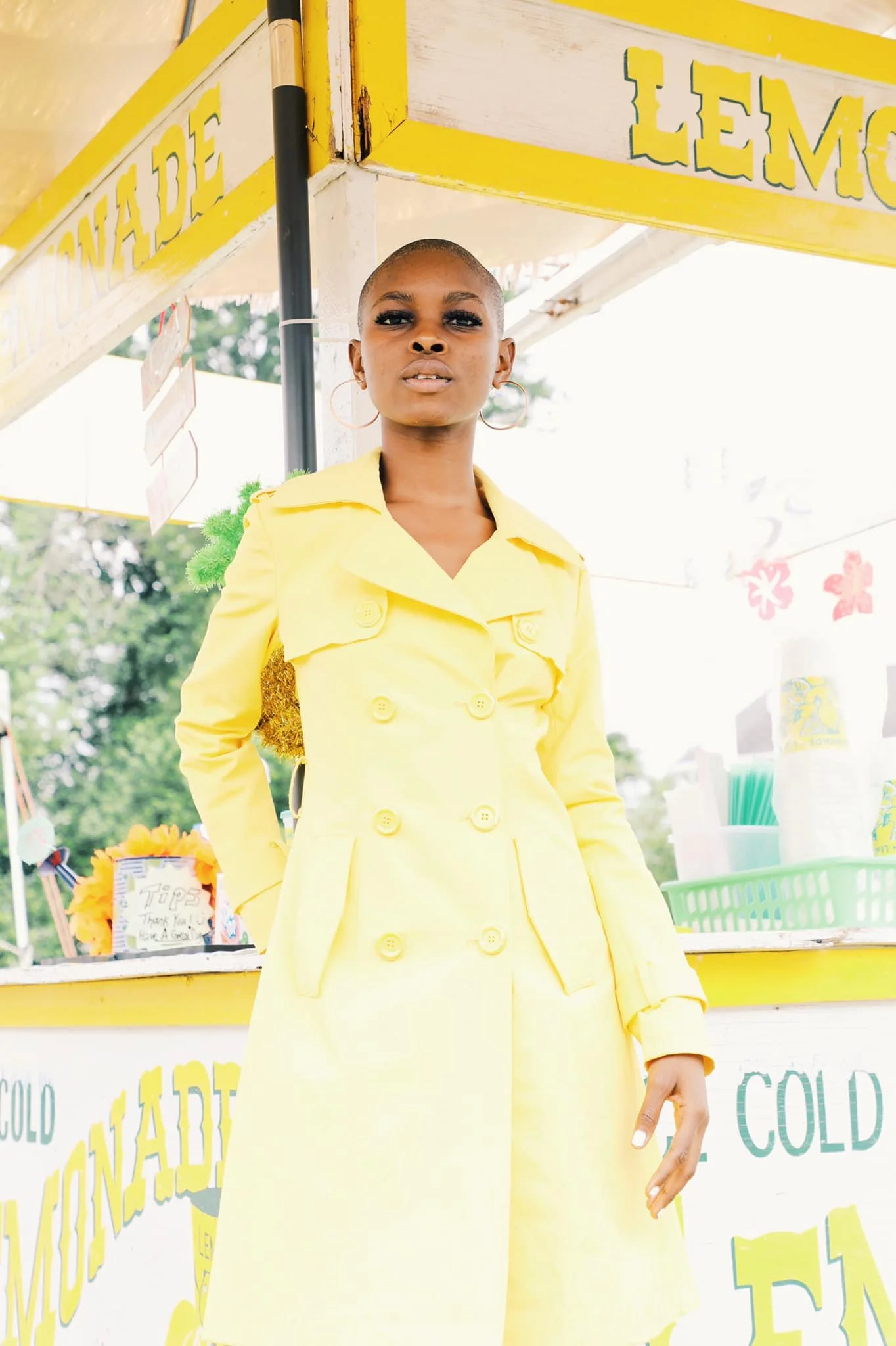 Woman in a yellow double-breasted trench coat standing in front of a lemon stand.