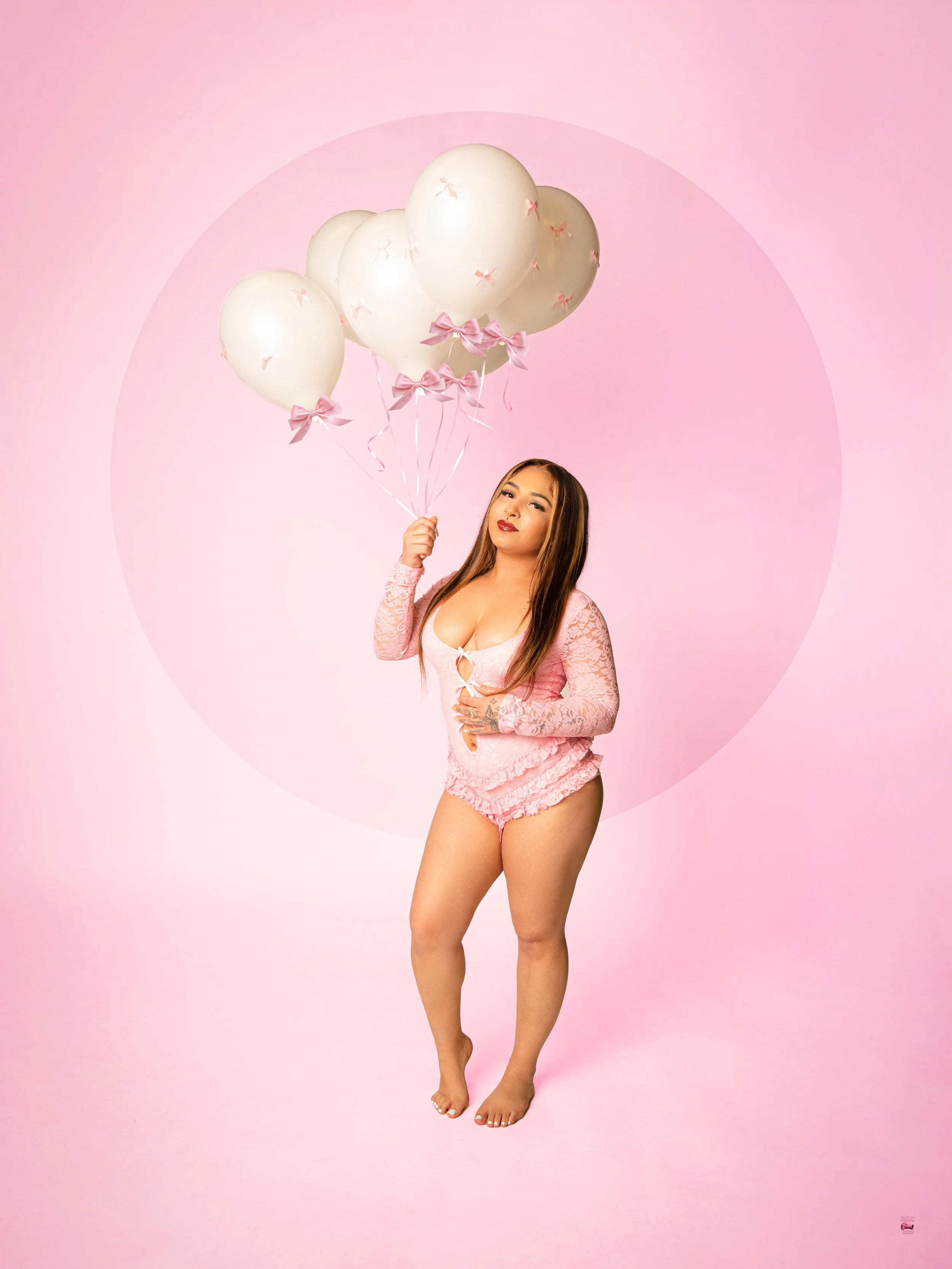 A woman in pink lace lingerie holding a bunch of white balloons with pink bows, standing against a pink background.