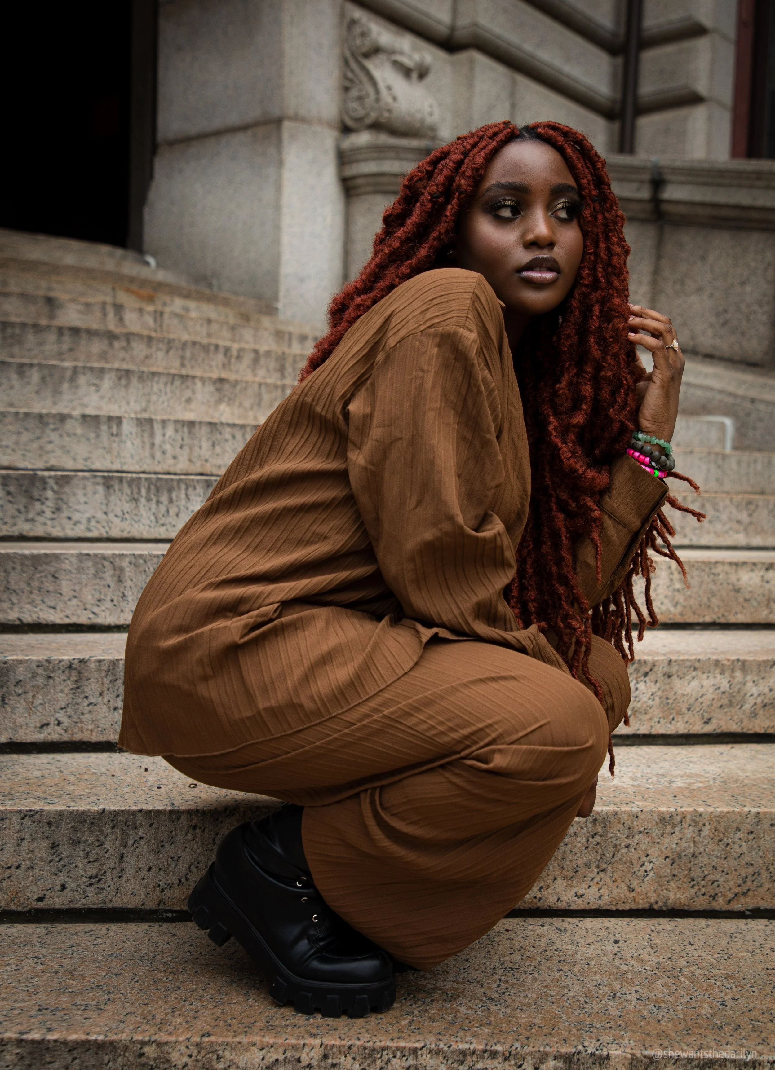 A woman with long red braided hair crouching on stone steps outside a building, wearing a brown outfit, black boots, and colorful bracelets, looking to the side.