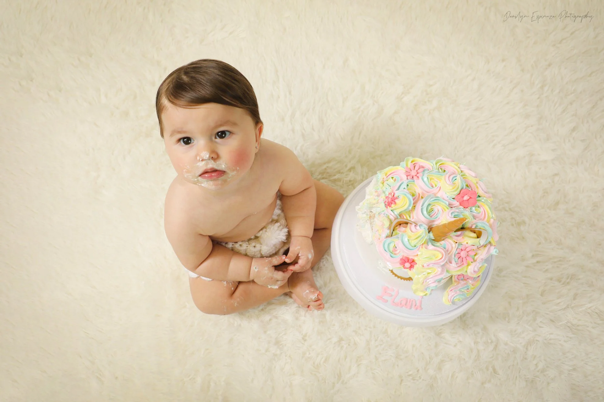 A baby sitting on a cream-colored fluffy rug next to a decorated birthday cake with pastel swirls and pink flowers.