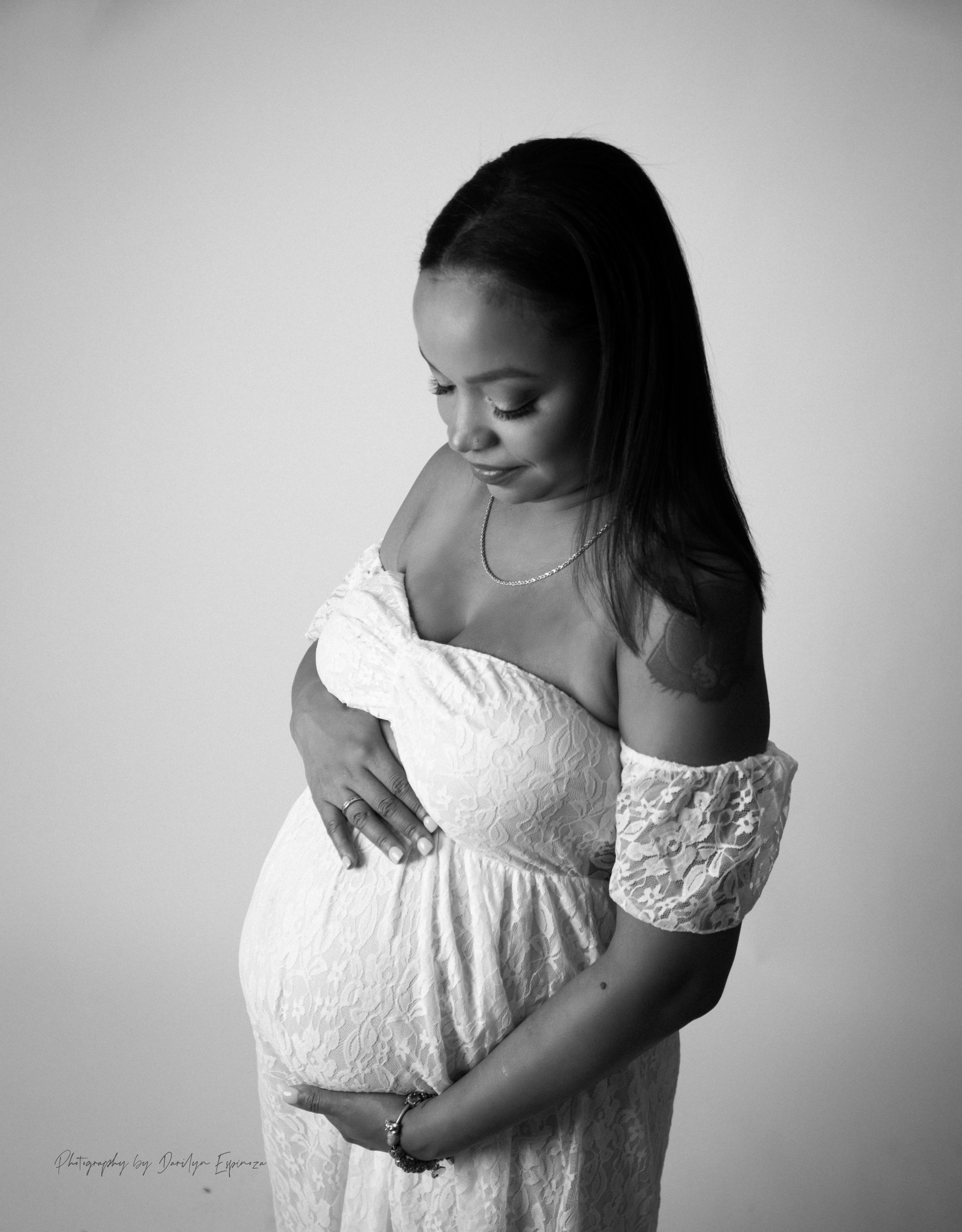 Black and white portrait of a pregnant woman wearing a off-the-shoulder lace dress, gently holding her pregnant belly, looking down with a serene expression.