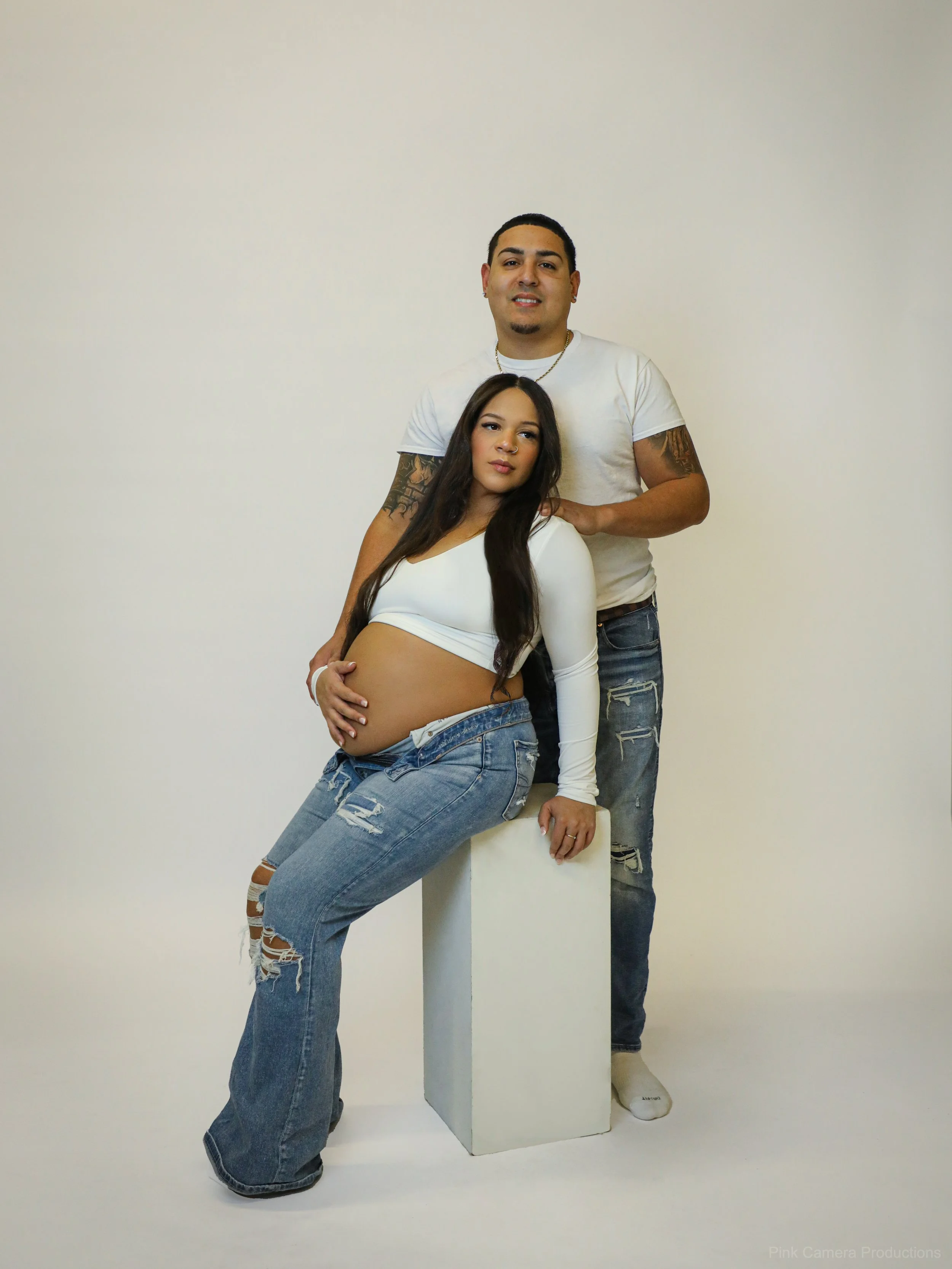 A pregnant woman sitting on a white podium with a man standing behind her, both wearing casual white tops and ripped jeans, against a plain white background.