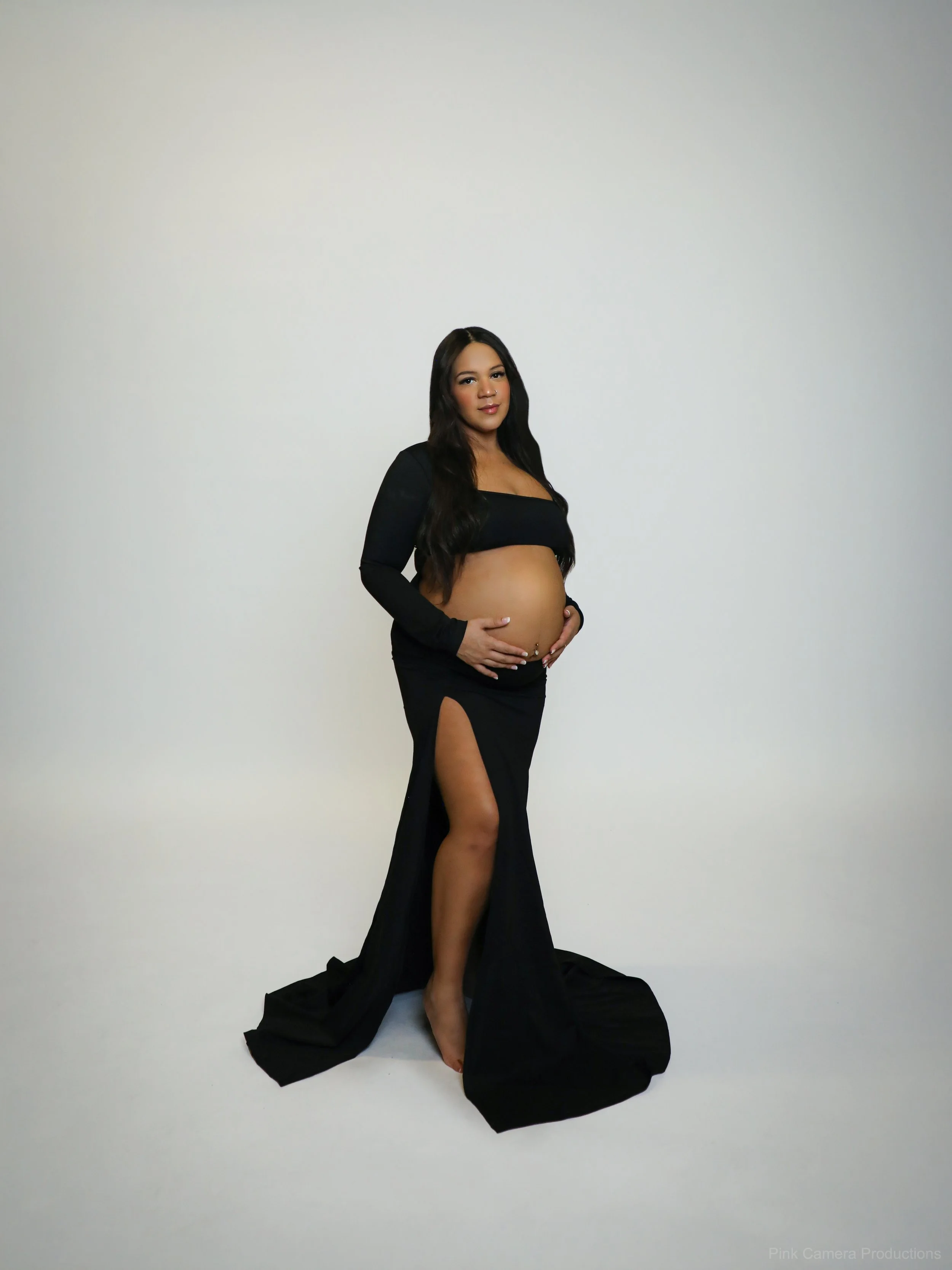 Pregnant woman standing in a black dress with a high slit, posing barefoot in front of a plain white background.