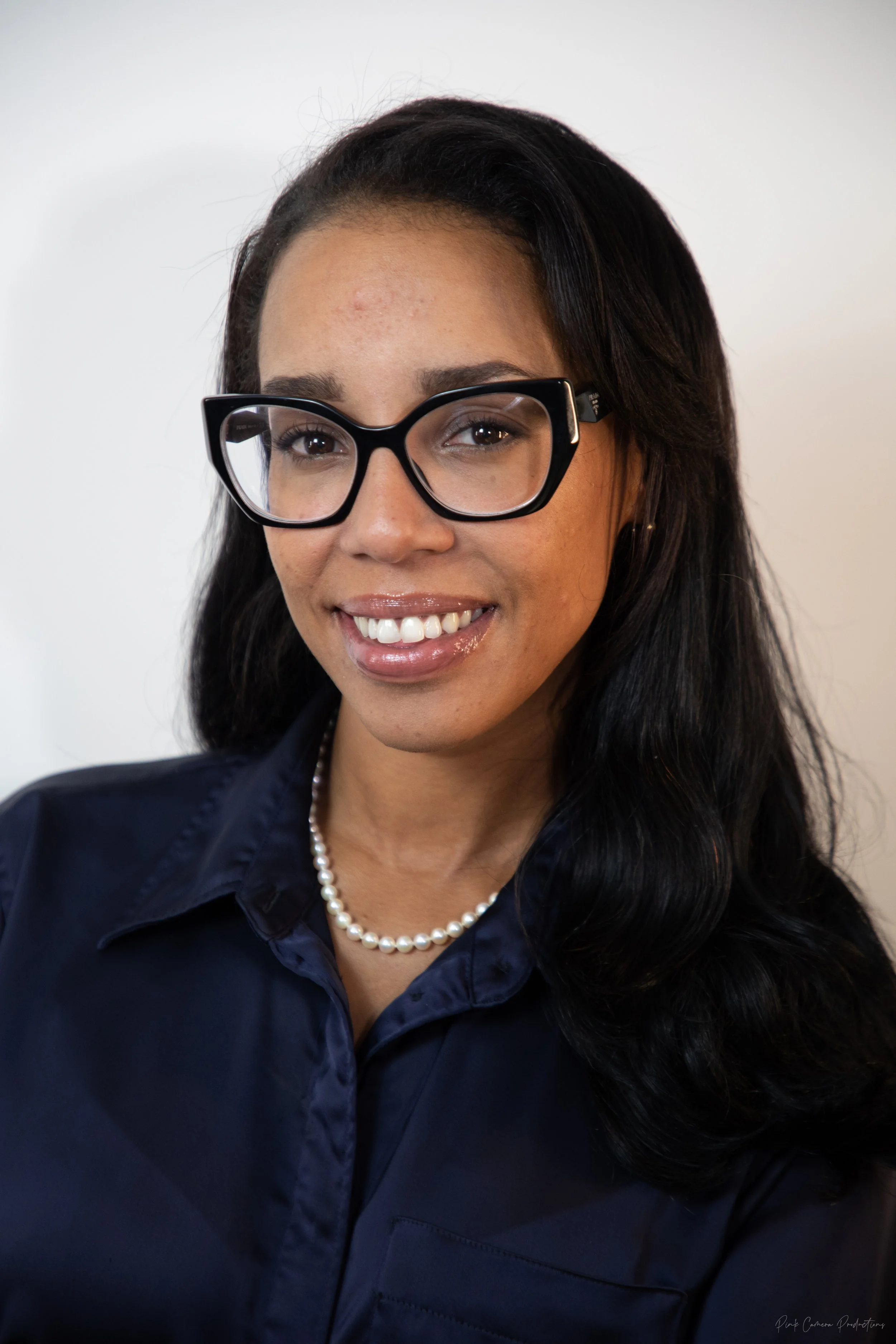 A woman wearing black glasses, a pearl necklace, and a dark blue shirt, smiling in front of a plain white background.