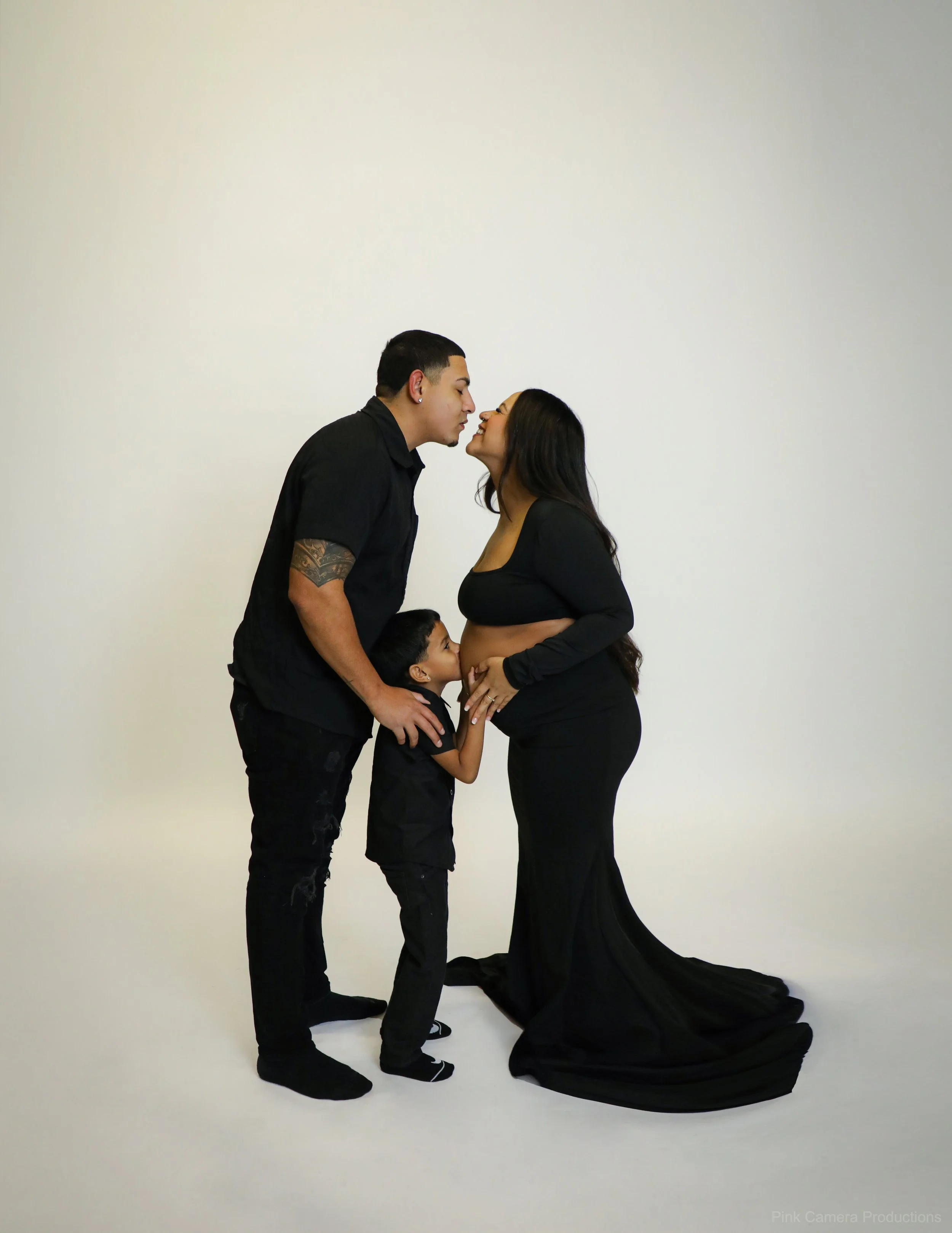 A family of three, a father, mother, and young child, sharing a close moment. The father and mother are about to kiss, while the child, a boy, touches the mother's pregnant belly. They are all dressed in black and are standing against a plain white b