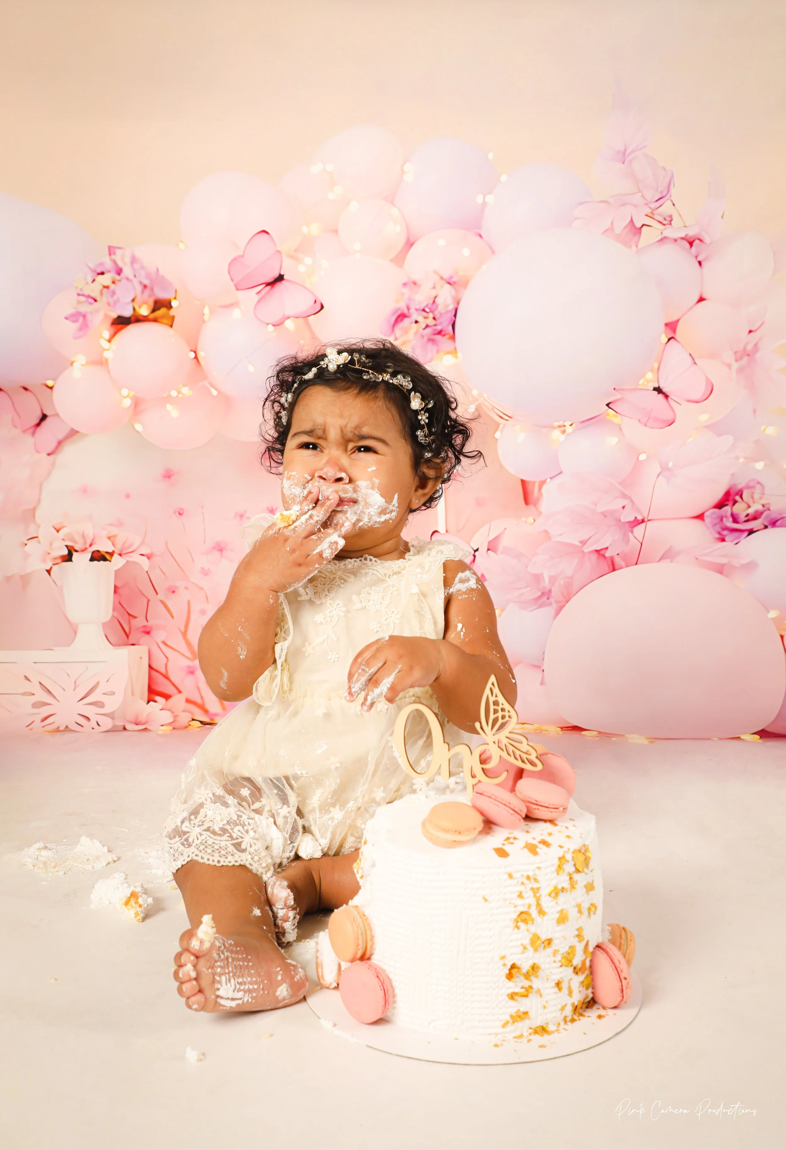 A young girl with curly hair, wearing a cream dress, sitting on the floor during her first birthday celebration. She has cake and frosting on her face and hands, with a surprised or playful expression. There is a birthday cake with pink macarons and 