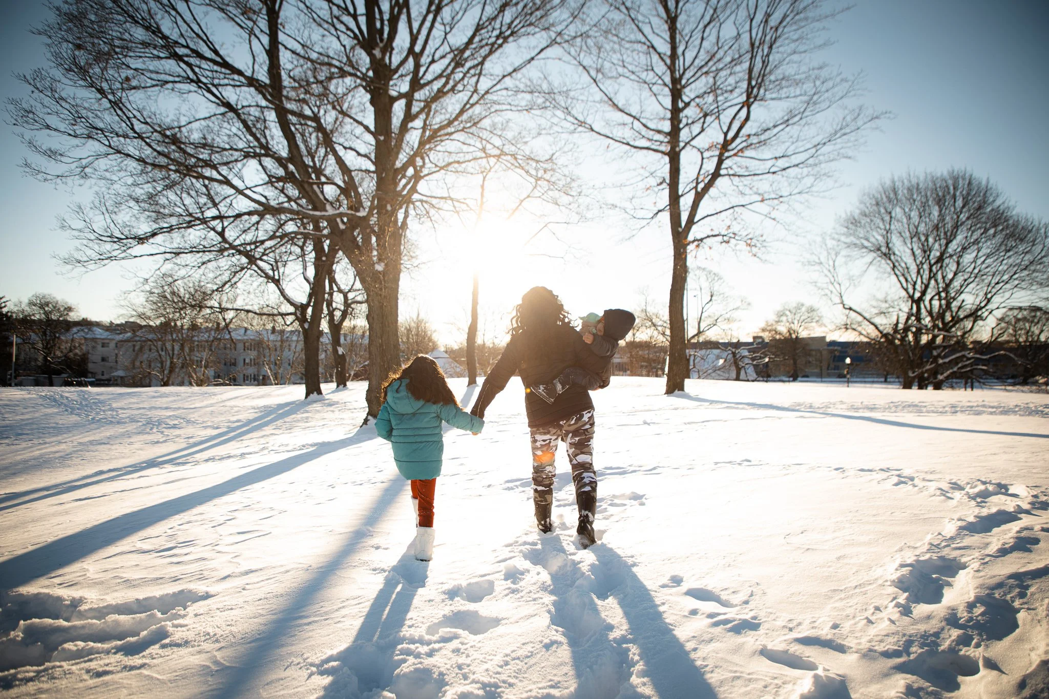 A woman and two children walking hand in hand through a snowy park on a sunny winter day, with leafless trees in the background.