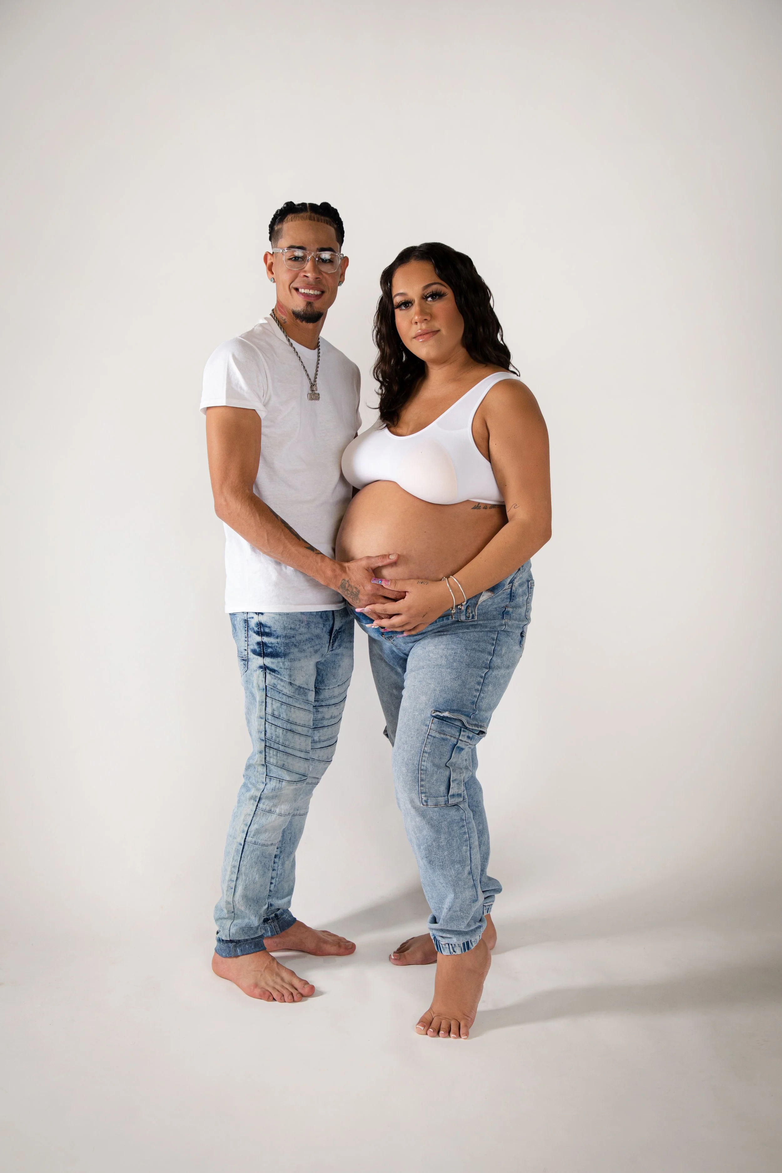 A pregnant woman and a man standing together, touching her pregnant belly, against a plain white background.