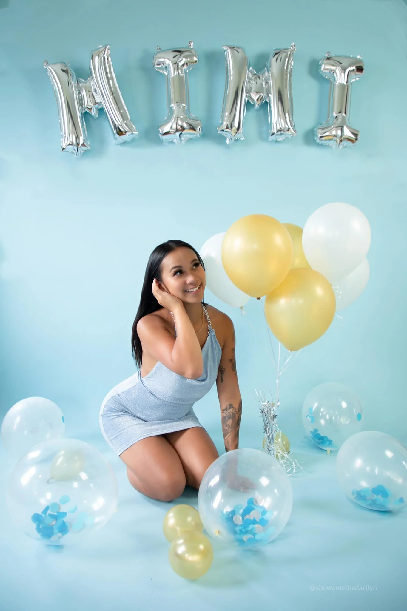 A woman kneeling on the floor at a birthday celebration with silver 'MINT' balloons on the wall behind her and a bunch of gold and white balloons around her. She is smiling and looking to the side.
