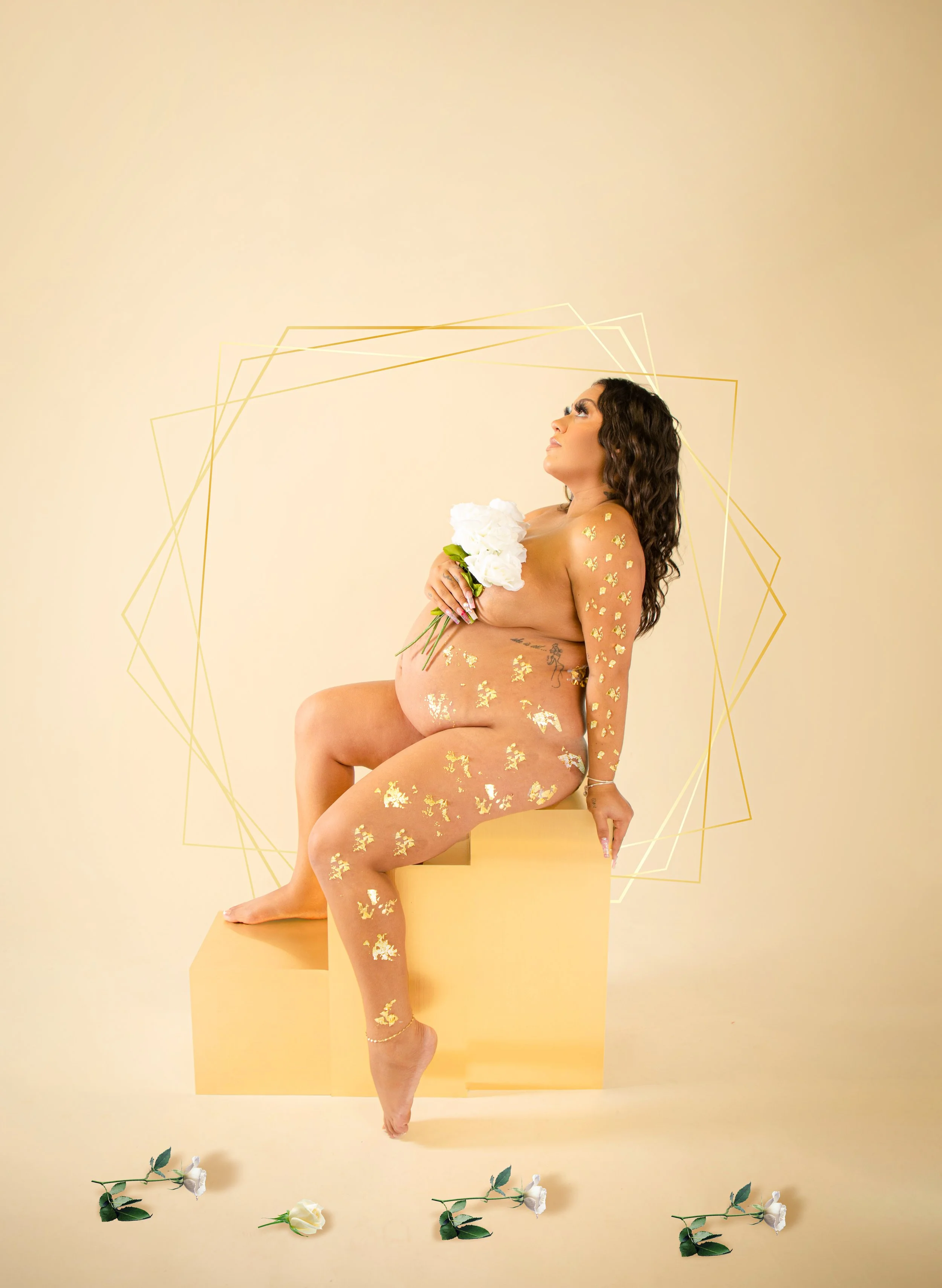 A nude woman with gold flower stickers sitting on a yellow block, holding white flowers, with her eyes looking up, gold geometric shapes behind her, and white roses on the floor.