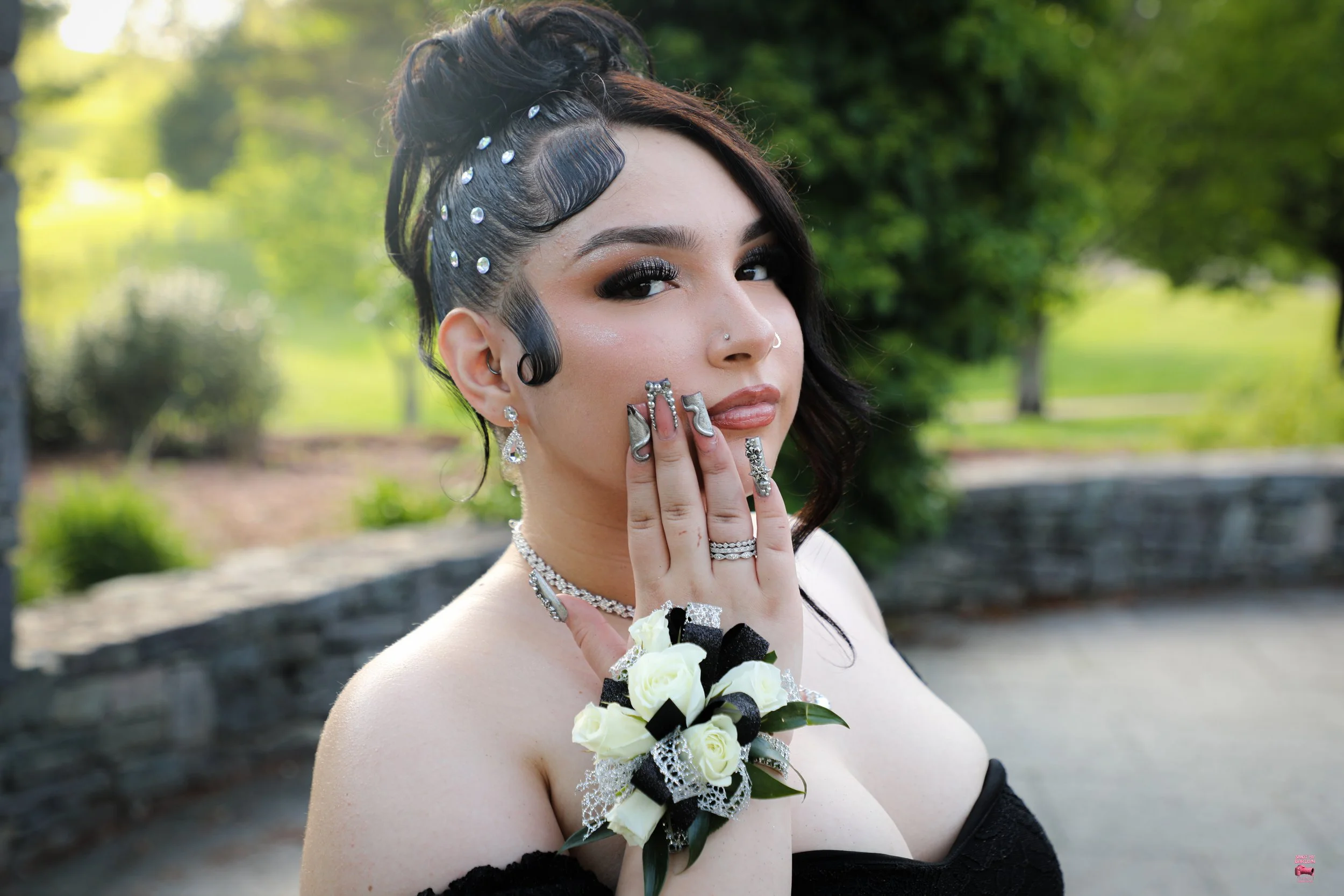 Portrait of a woman with elaborate black and pearl hair styling, wearing multiple rings, earrings, and a choker, holding her hand near her face, outdoors with green trees in the background.