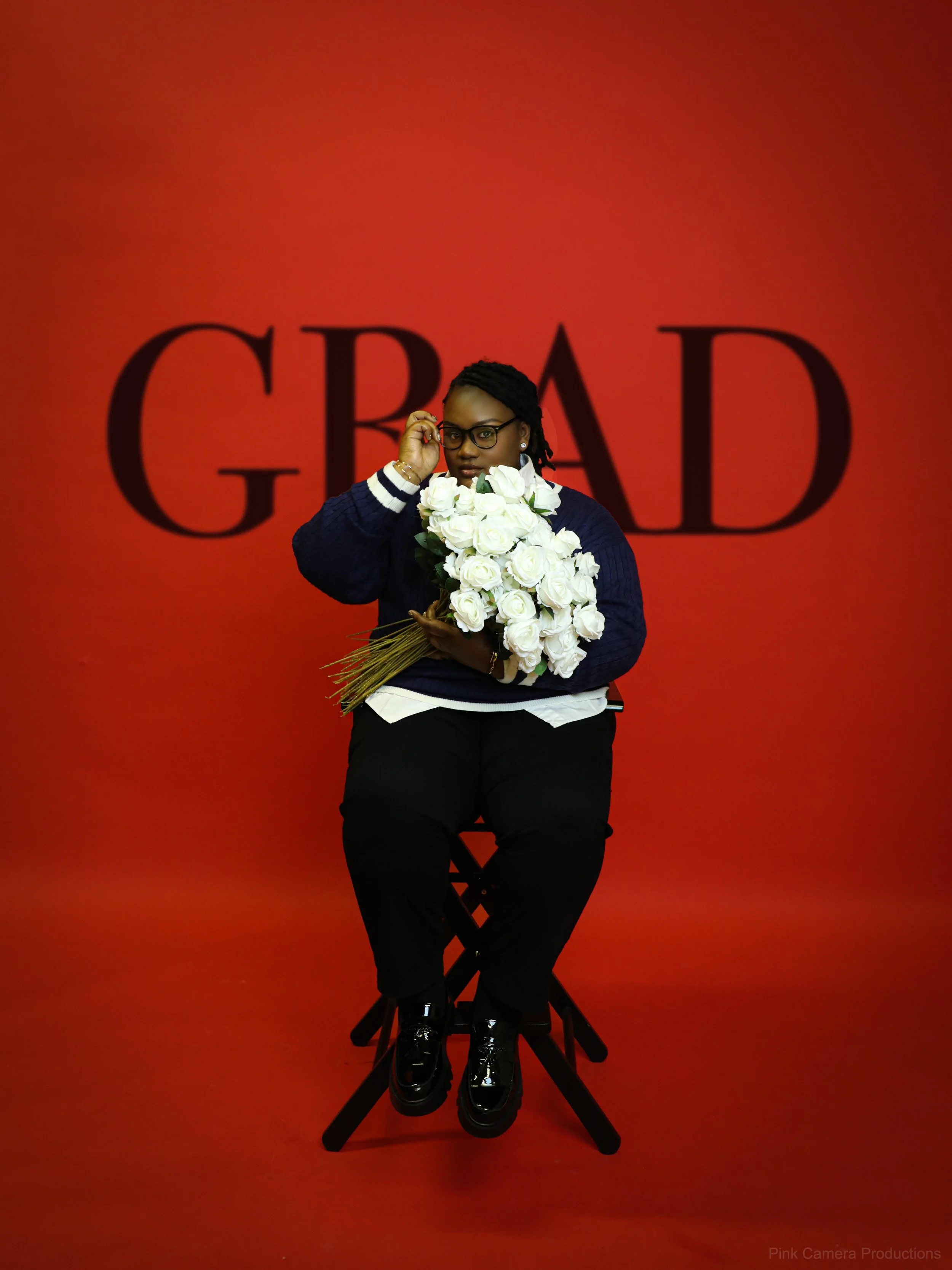 Woman sitting on a stool holding a large bouquet of white roses, wearing glasses and a dark sweater, with a large word 'GREAT' in the background on a red wall.