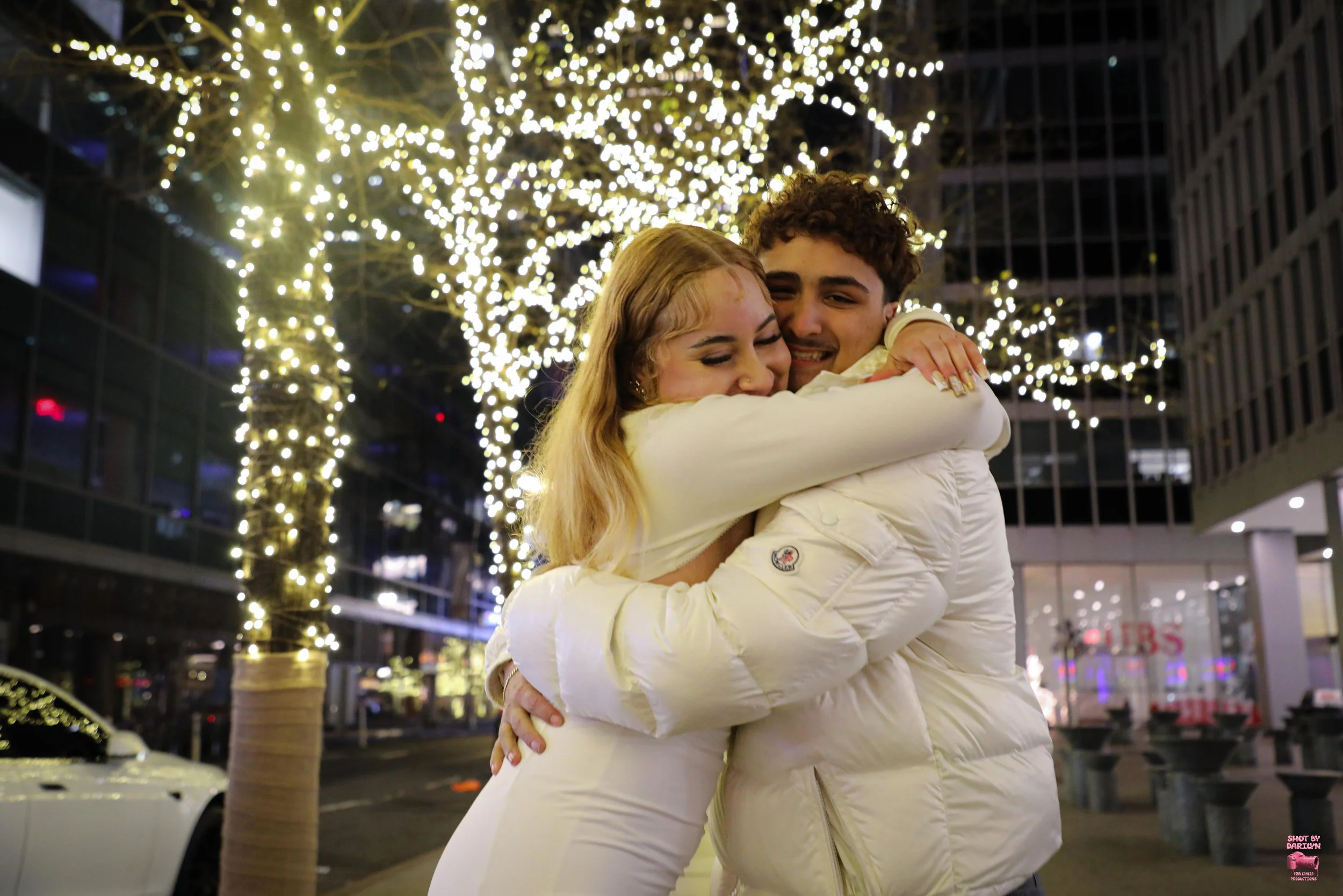 A young couple embracing and smiling on a city street decorated with twinkling holiday lights on trees at night.