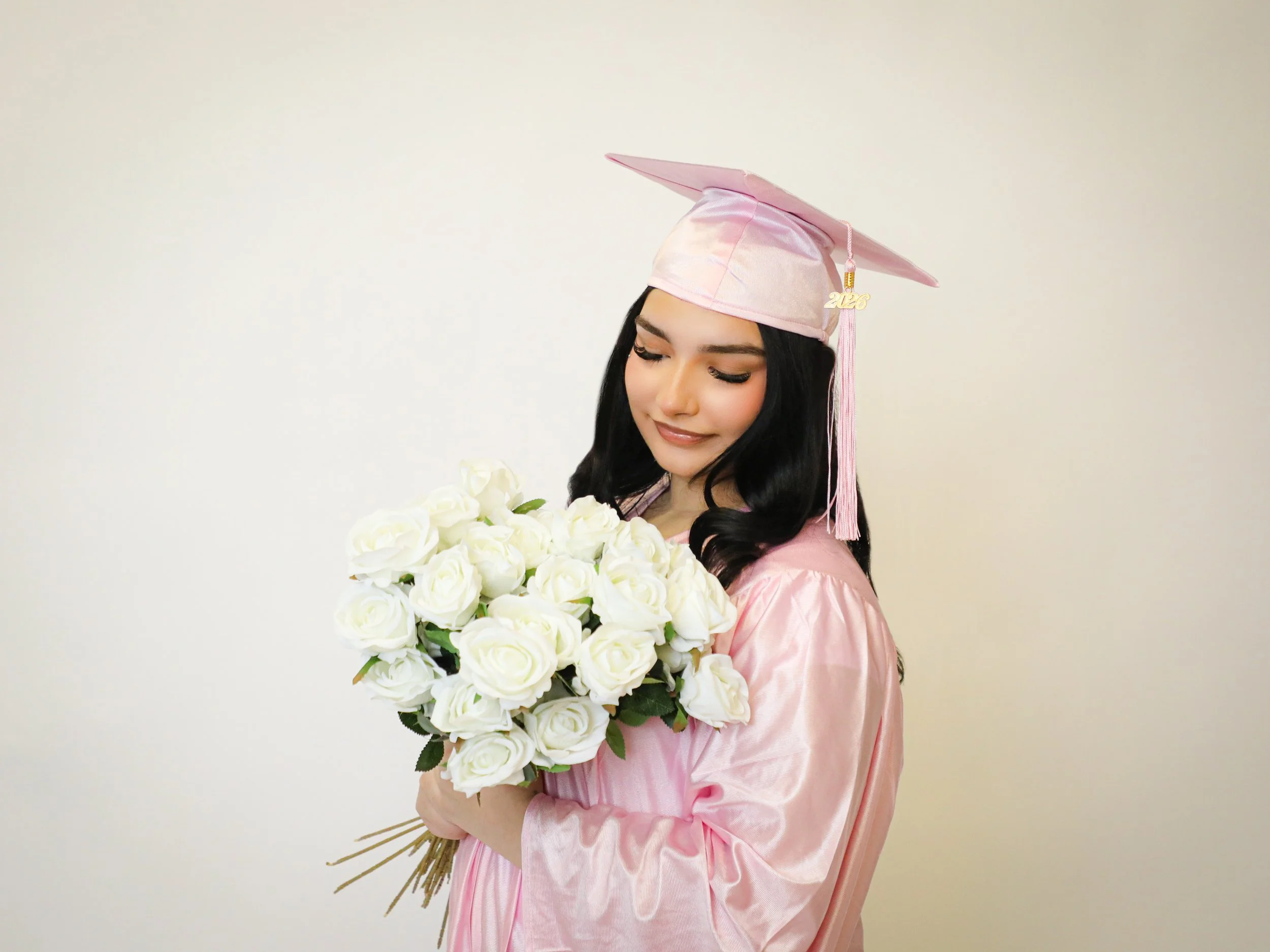 A young woman in a pink graduation gown and cap holding a bouquet of white roses, smiling with her eyes closed.